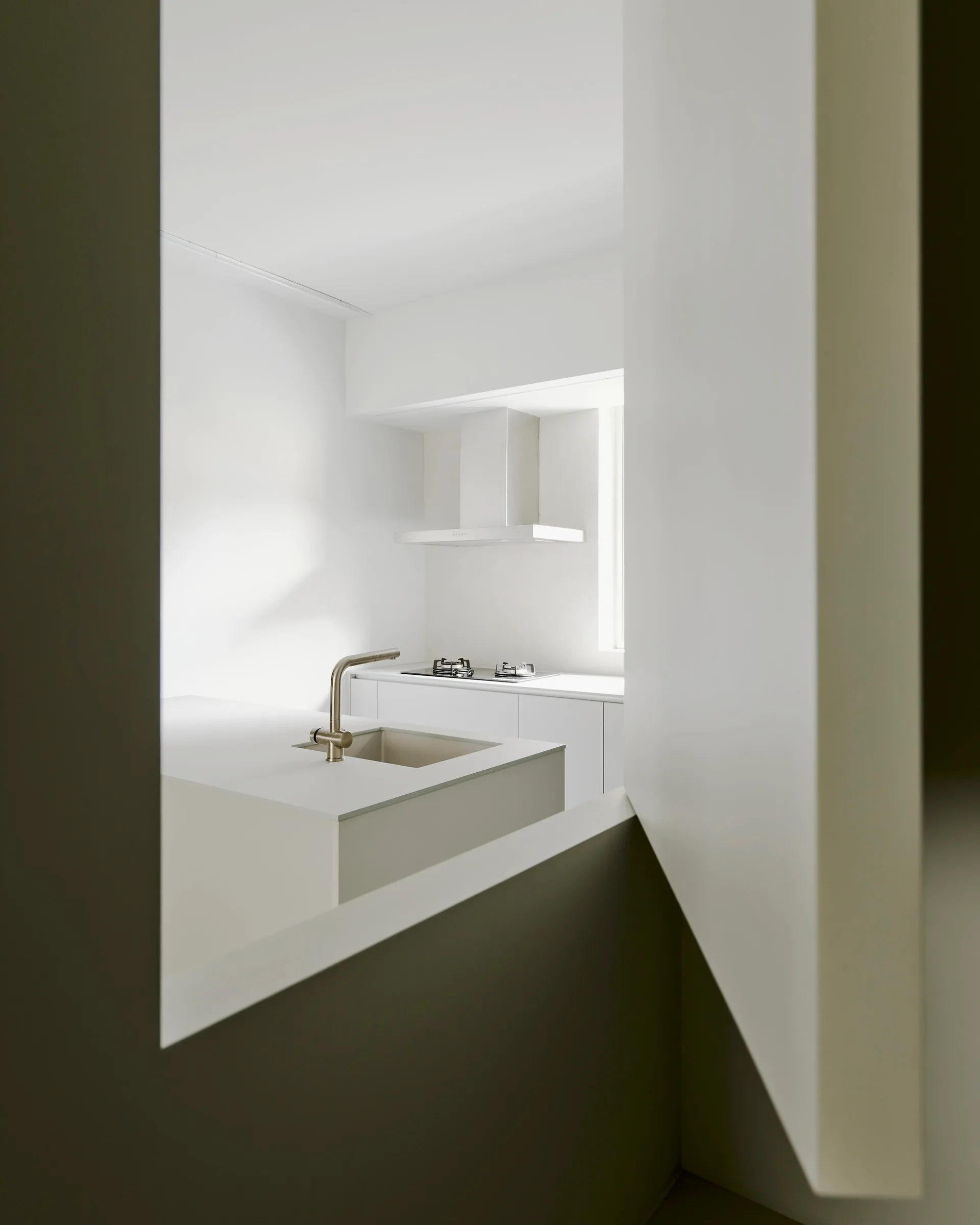 A view through a wall opening reveals a minimalist white kitchen featuring a central island with a metal faucet, integrated cabinetry, and a cooktop.