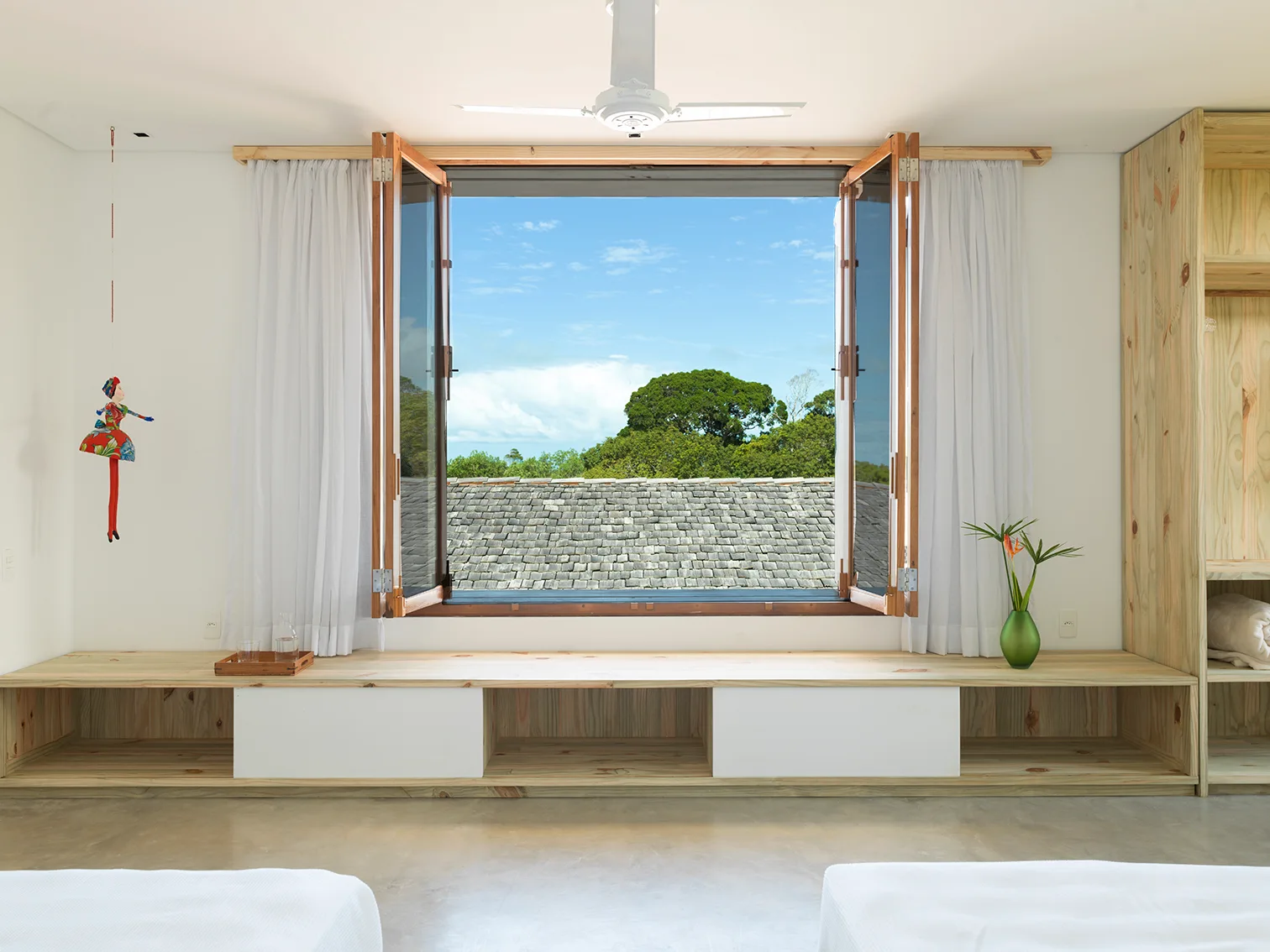 A bright bedroom features a large open wooden window and a low built-in pine bench that extends across the wall below white sheer curtains.