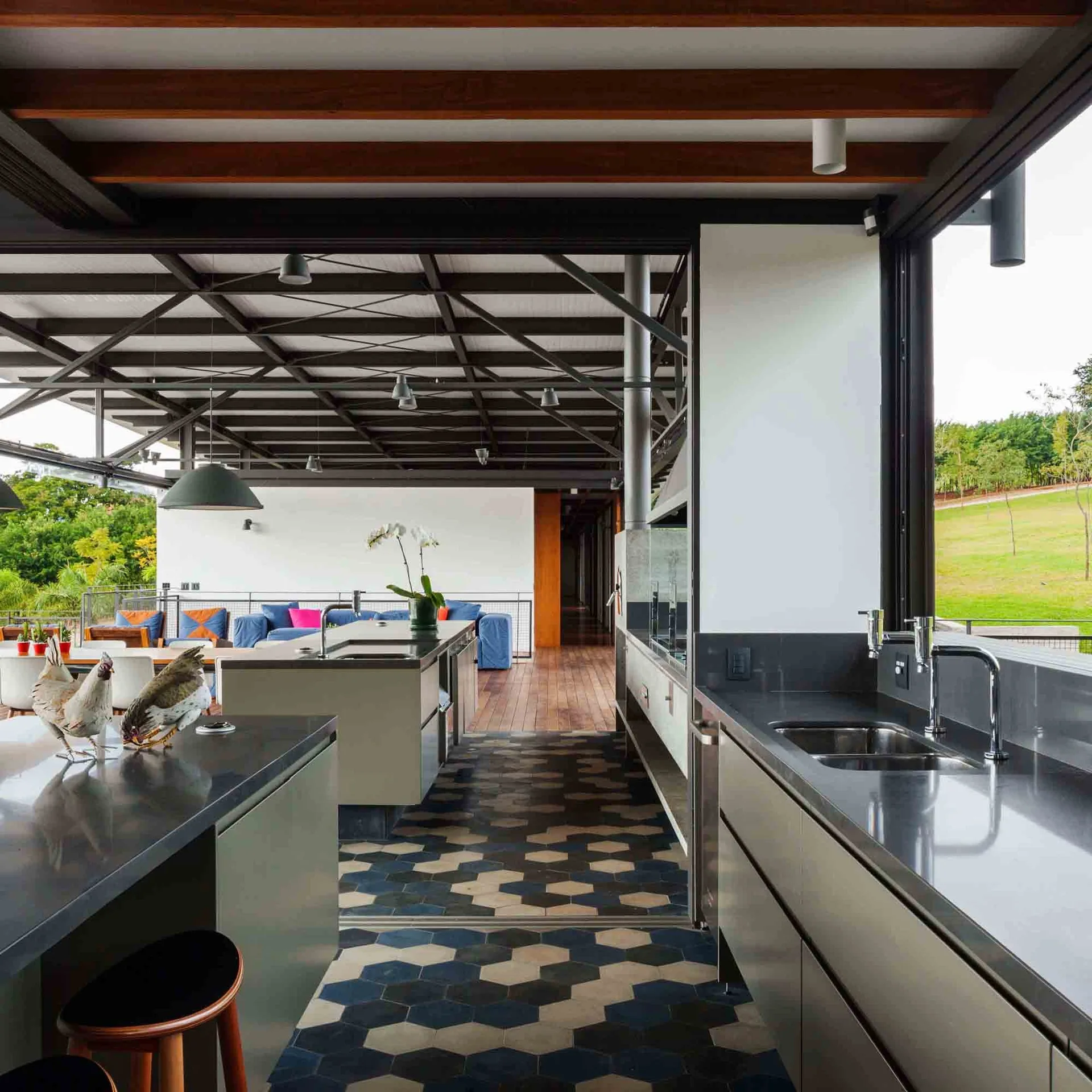 A modern open-plan kitchen features dark grey countertops, patterned blue and grey hexagonal floor tiles, and an industrial metal truss ceiling.