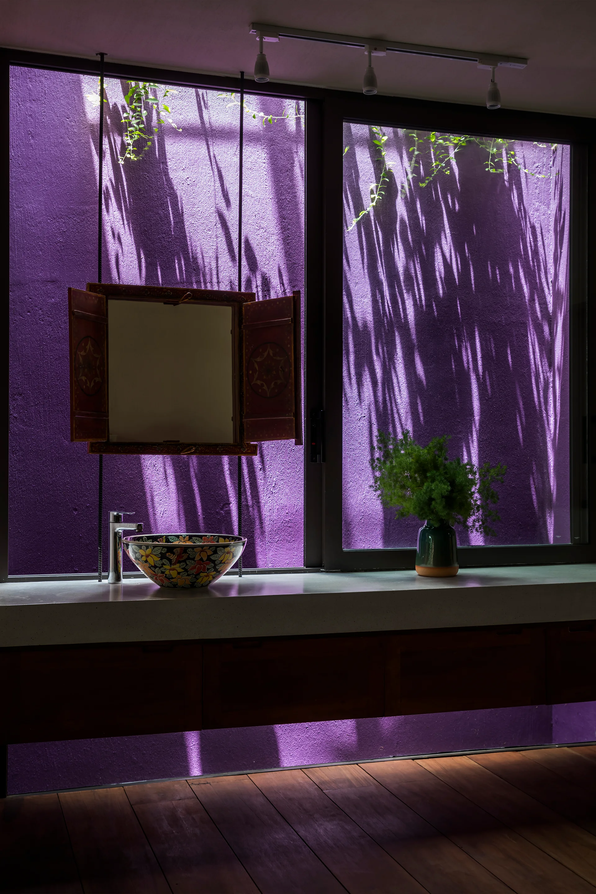 A bathroom features a concrete countertop with a colorful vessel sink and a shuttered wooden mirror set against a window looking out at a purple wall.