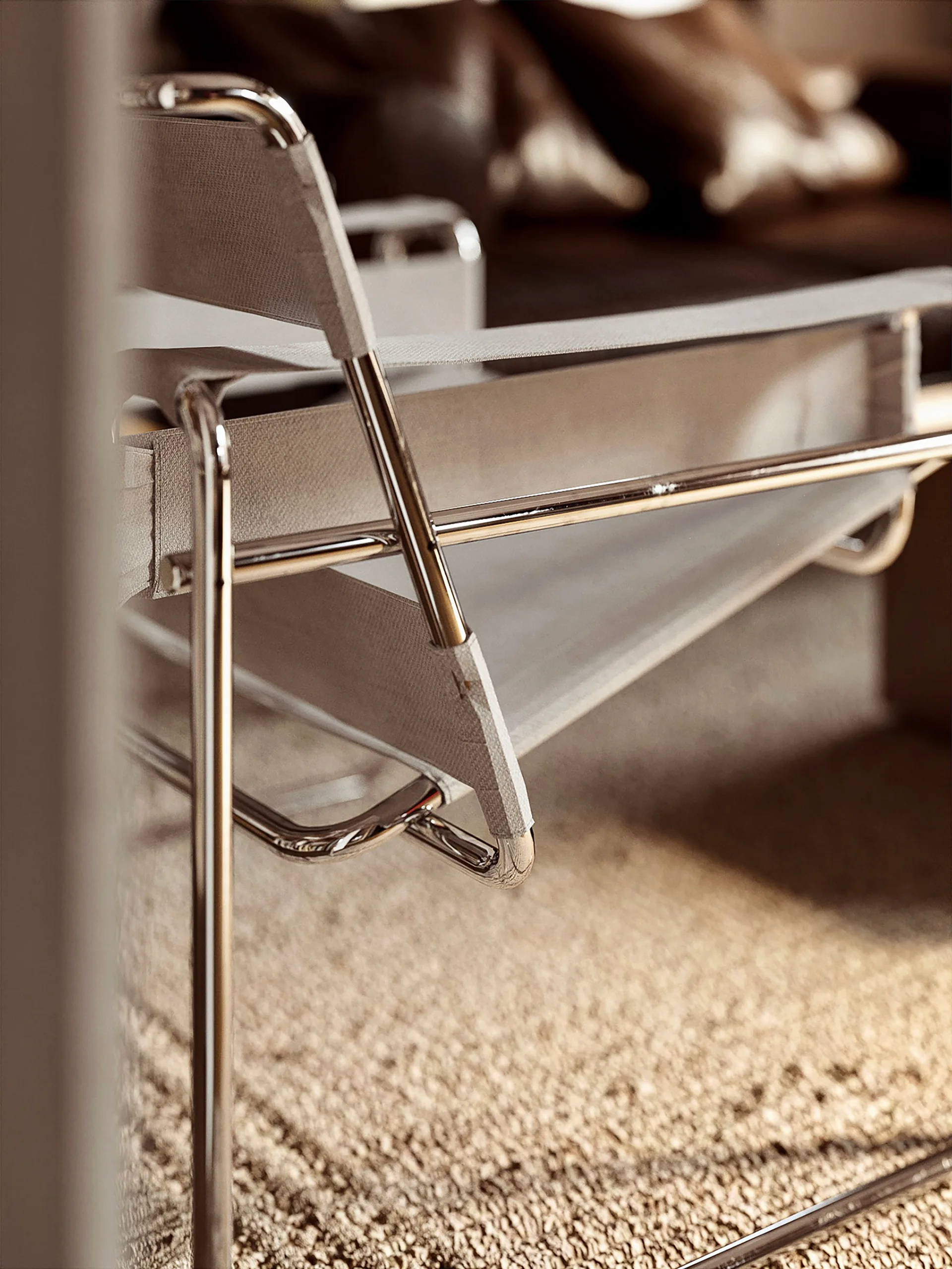 A close-up of a modern minimalist armchair featuring a polished chrome tubular frame and light beige canvas straps over a textured rug.