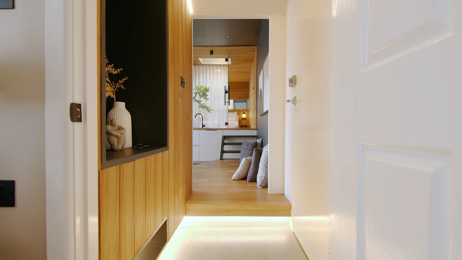 A transitional hallway featuring a wood-paneled wall with a black recessed display niche and an illuminated wood step leading into a room.