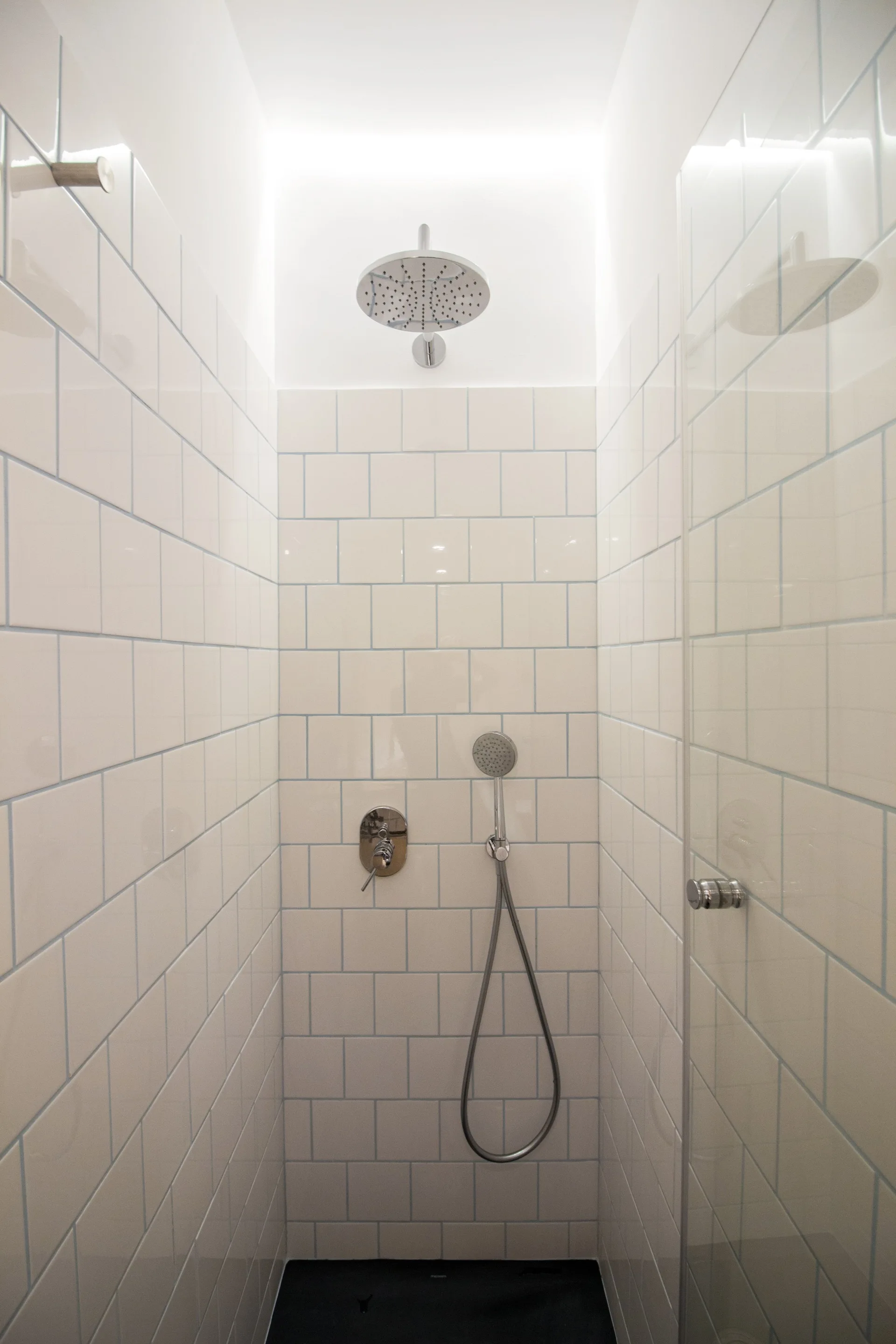 A narrow shower stall features white square ceramic wall tiles with blue grout lines, chrome fixtures, and a glass partition.