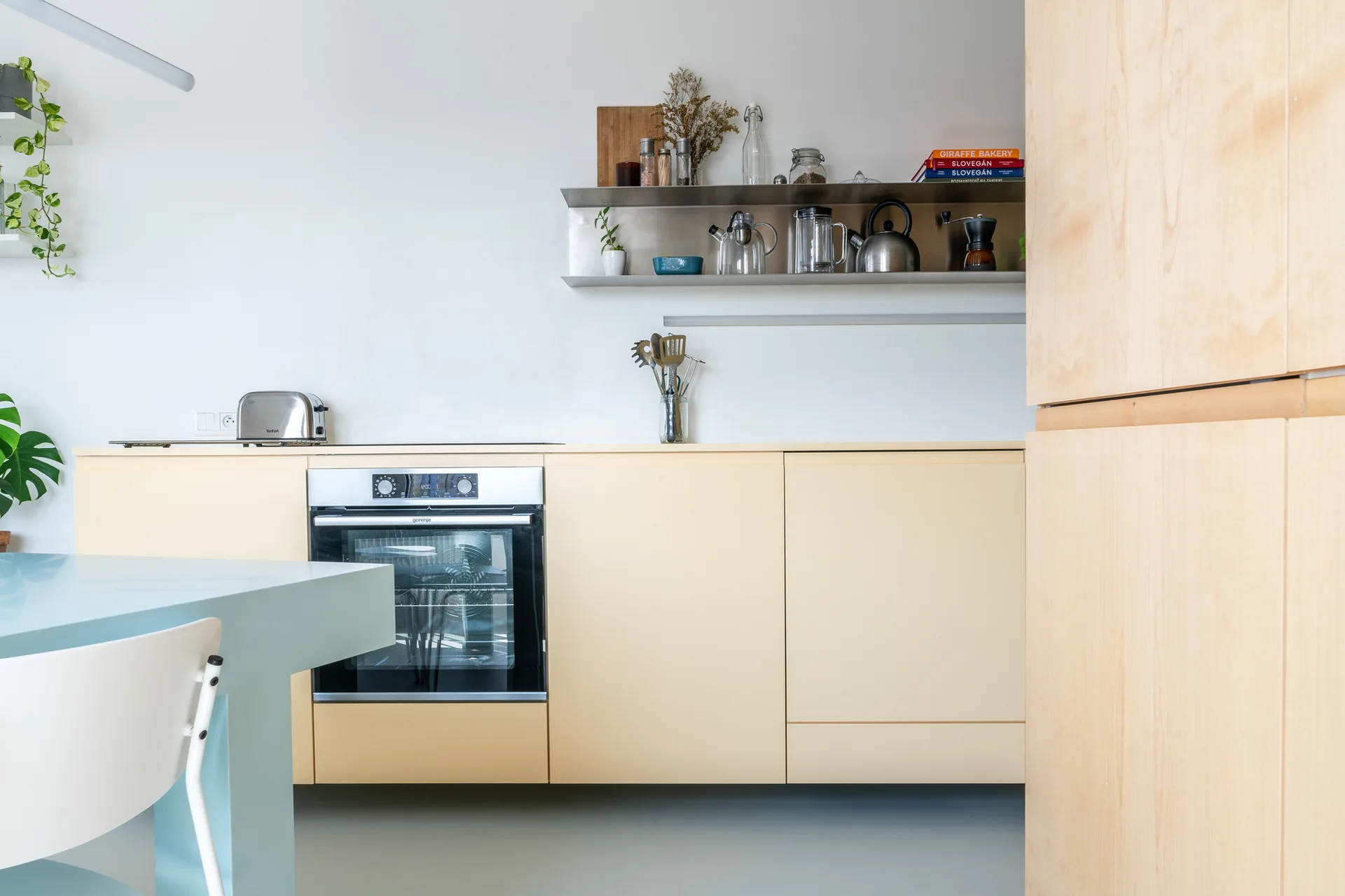 A minimalist kitchen featuring pale yellow flat-front cabinets, a stainless steel wall shelf with jars, and a light blue dining table.