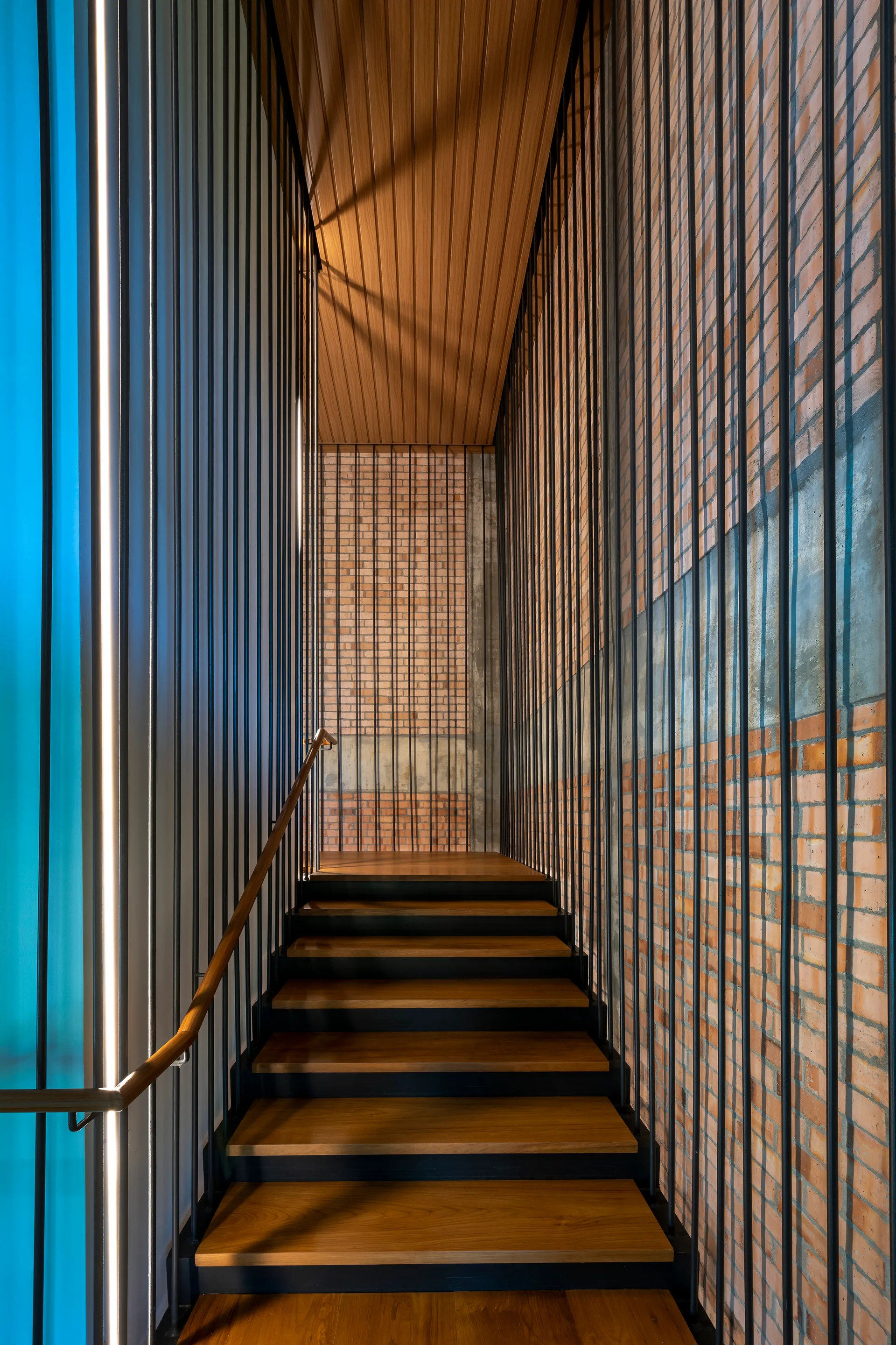 A modern industrial staircase features wooden treads, vertical black steel spindles, a slatted wood ceiling, and an exposed brick and concrete wall.