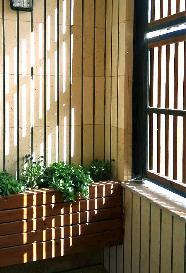 A wooden bench with green plants sits in a tiled corner next to a window with vertical wooden slats casting linear shadows.