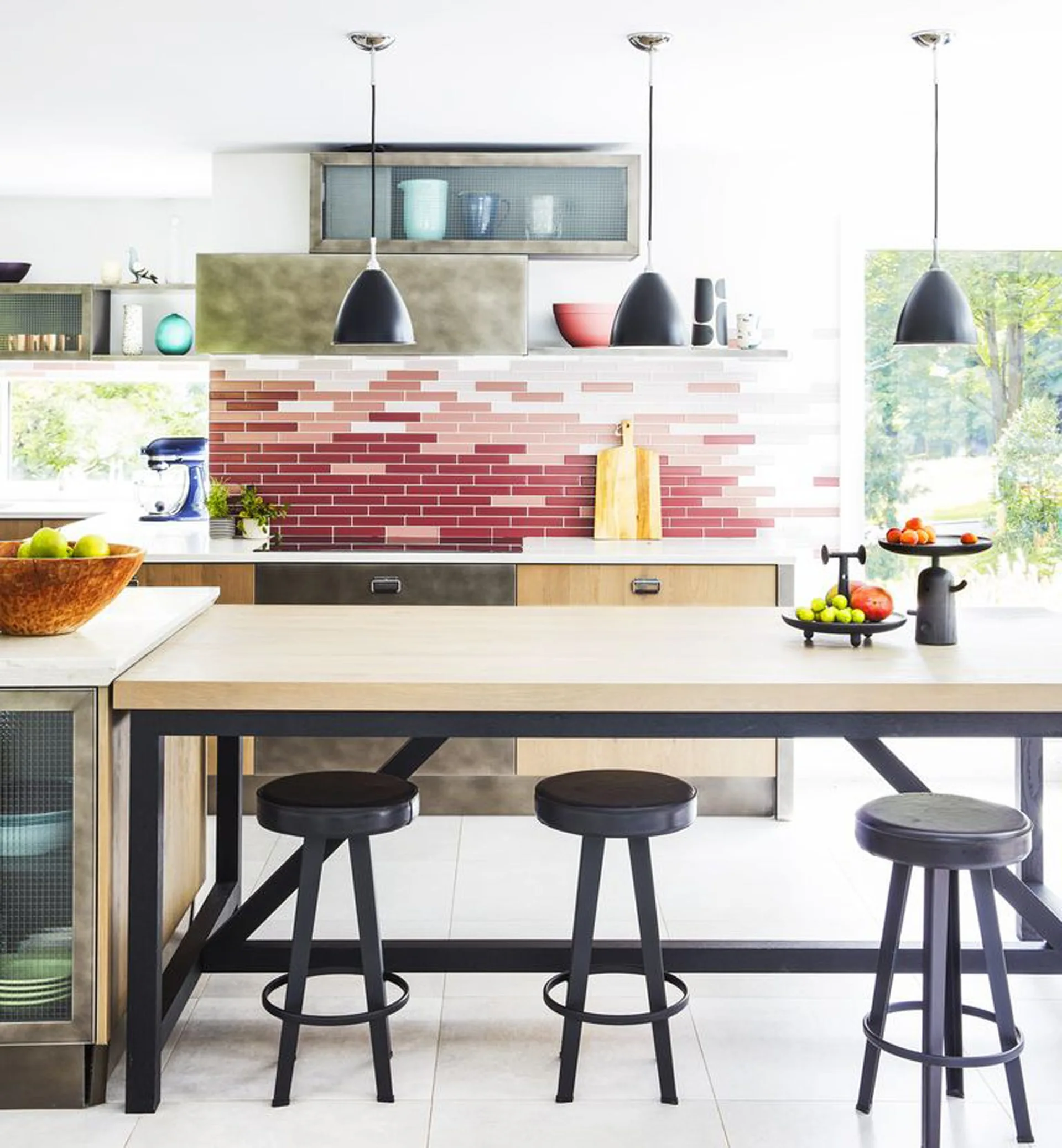 A bright contemporary kitchen features a red ombre tiled backsplash behind wood cabinetry and a large wooden island with three black metal stools.