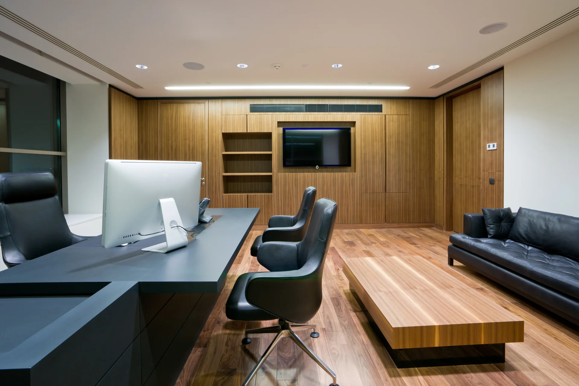 A modern office featuring a black desk with computer, black leather chairs, a matching sofa, and vertical wood paneling on the back wall.