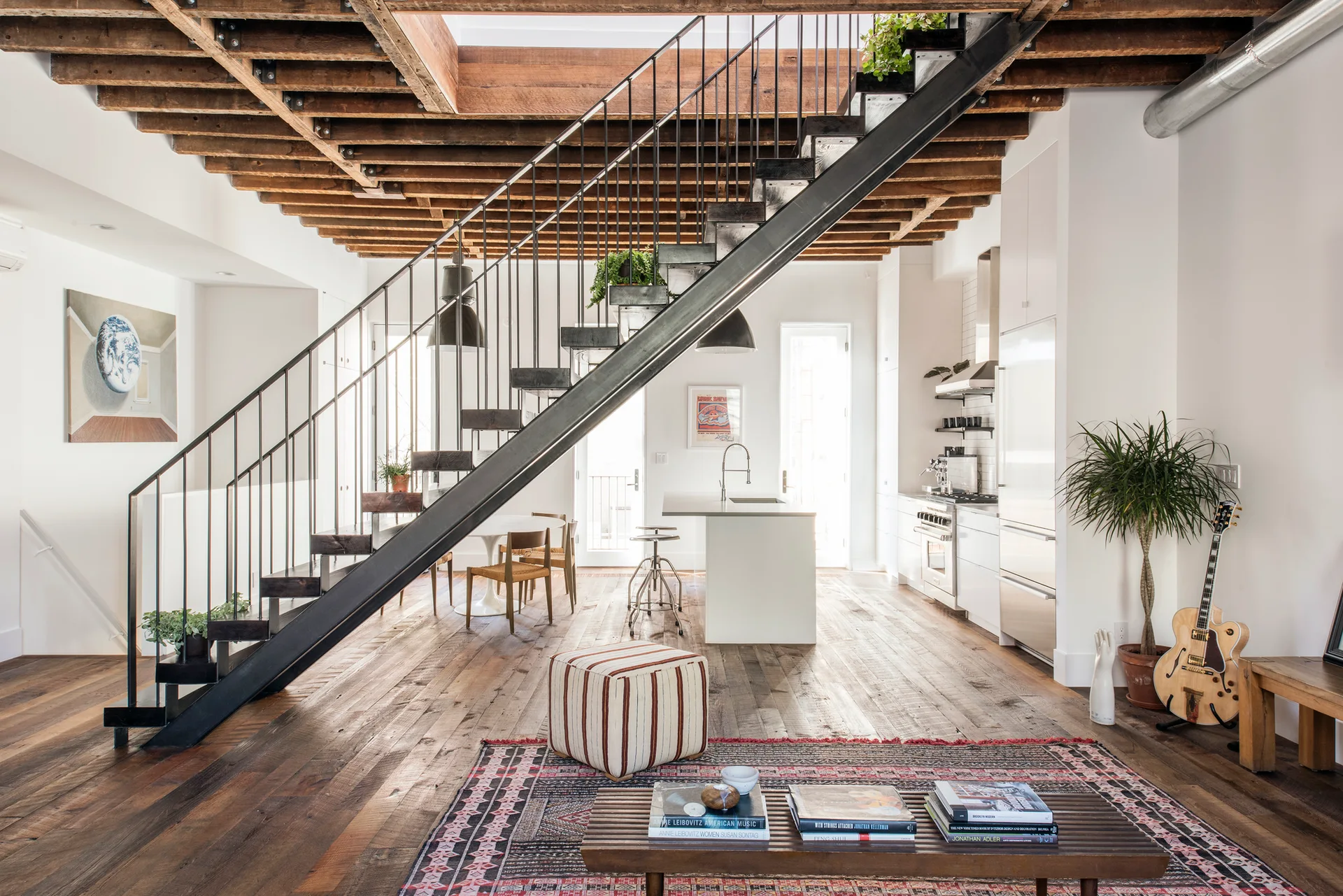 An open loft space features an industrial black metal staircase, exposed wooden ceiling joists, a white kitchen island, and reclaimed wood floors.