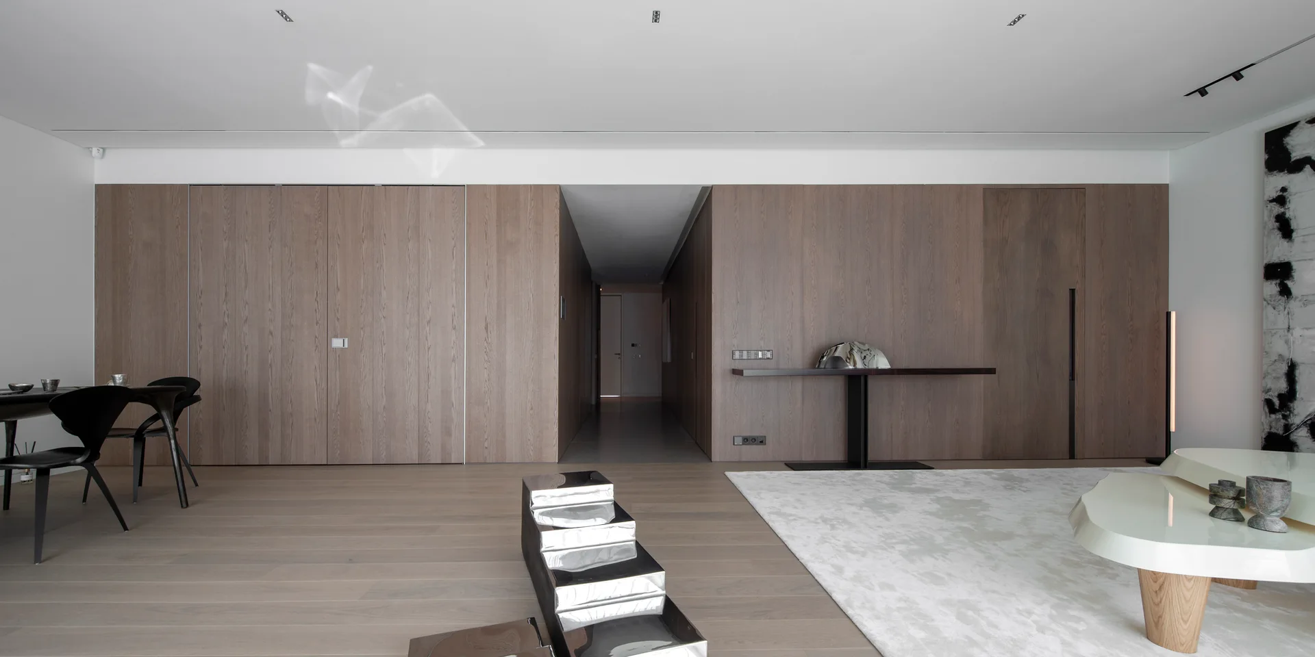 A minimalist living room features vertical wood wall paneling with integrated flush doors, light hardwood floors, and a sculptural cream coffee table.