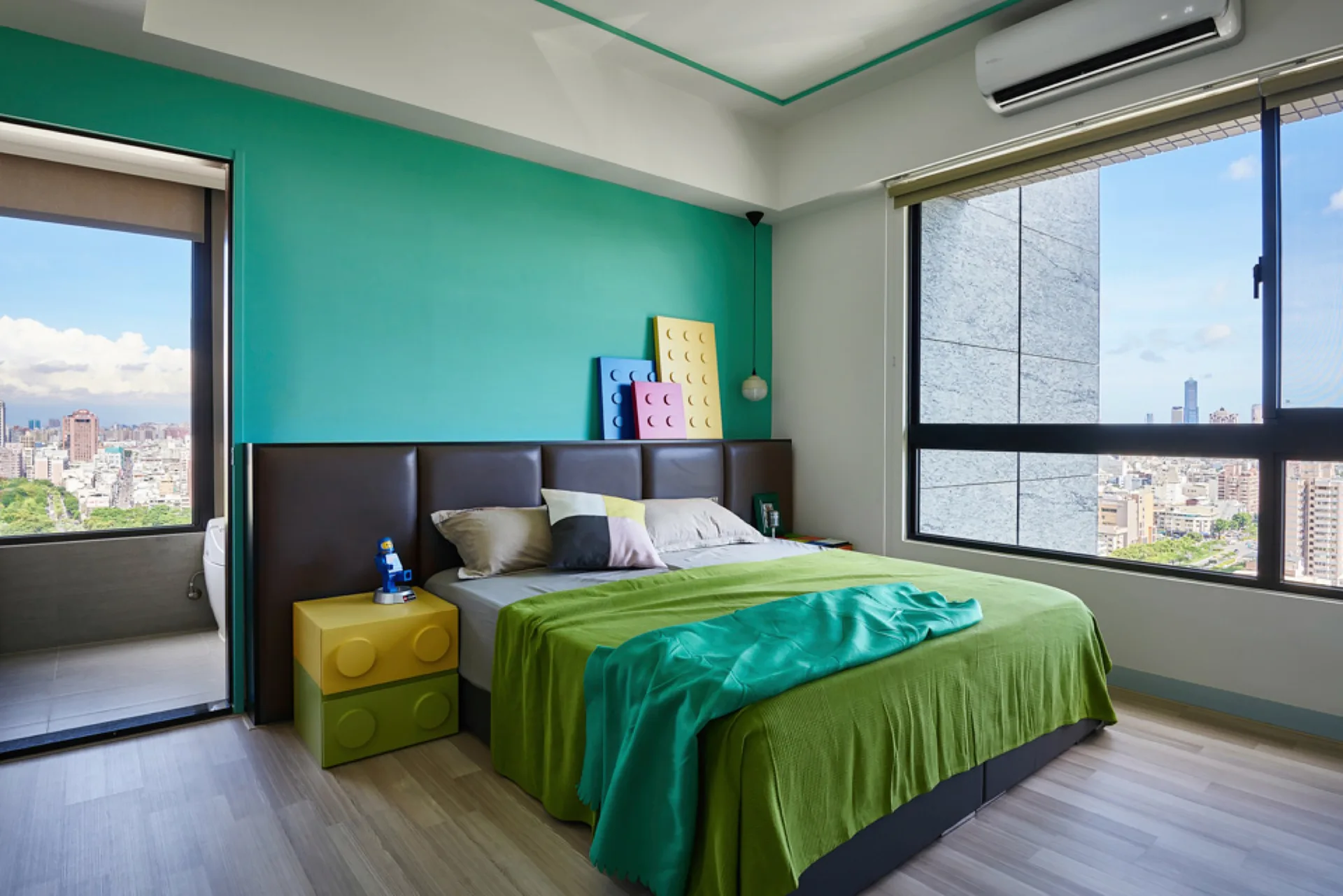A bedroom with a teal accent wall features a bed with green bedding, a brown leather headboard, and yellow and green block-shaped modular nightstands.