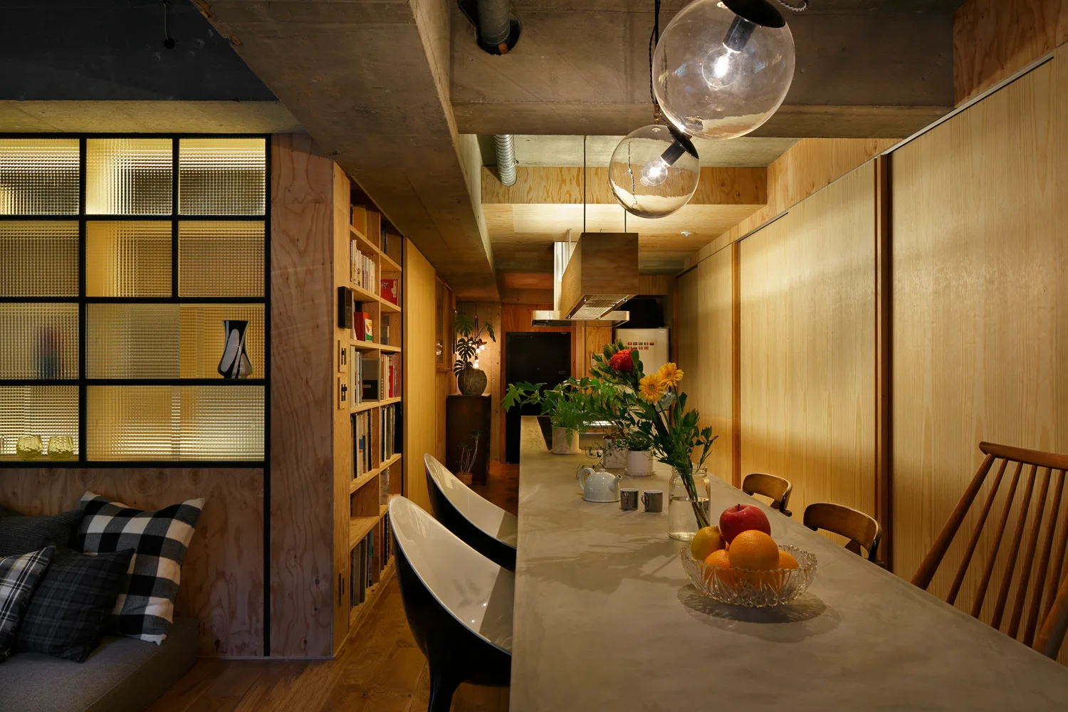 A long concrete kitchen island with bar stools sits beneath exposed beams next to a reeded glass partition and plywood walls.