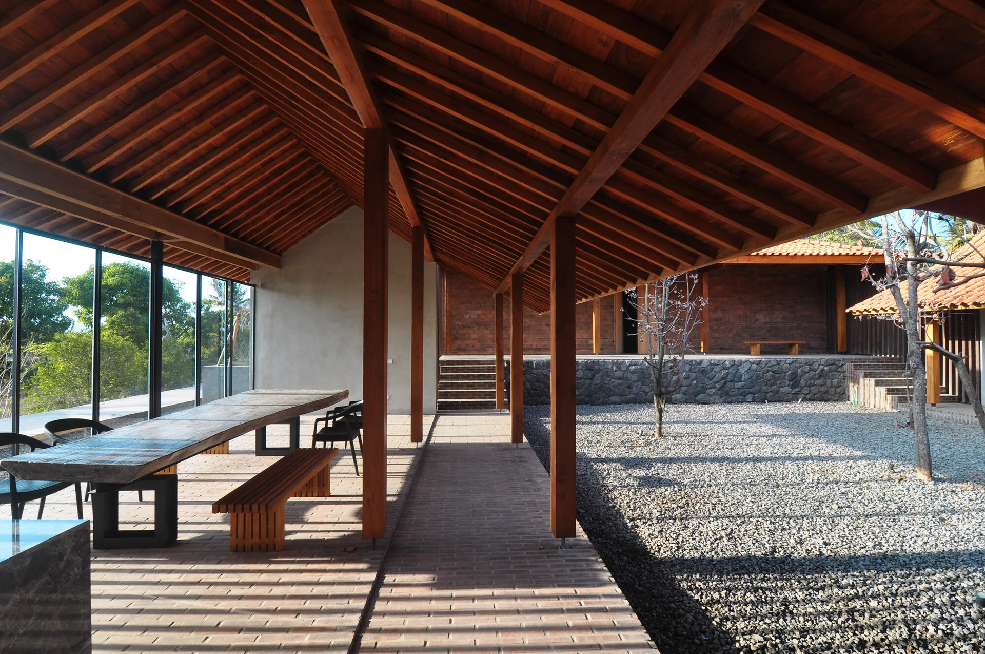 An open-air covered patio features a long wooden dining table and bench beneath a pitched roof with exposed timber beams.