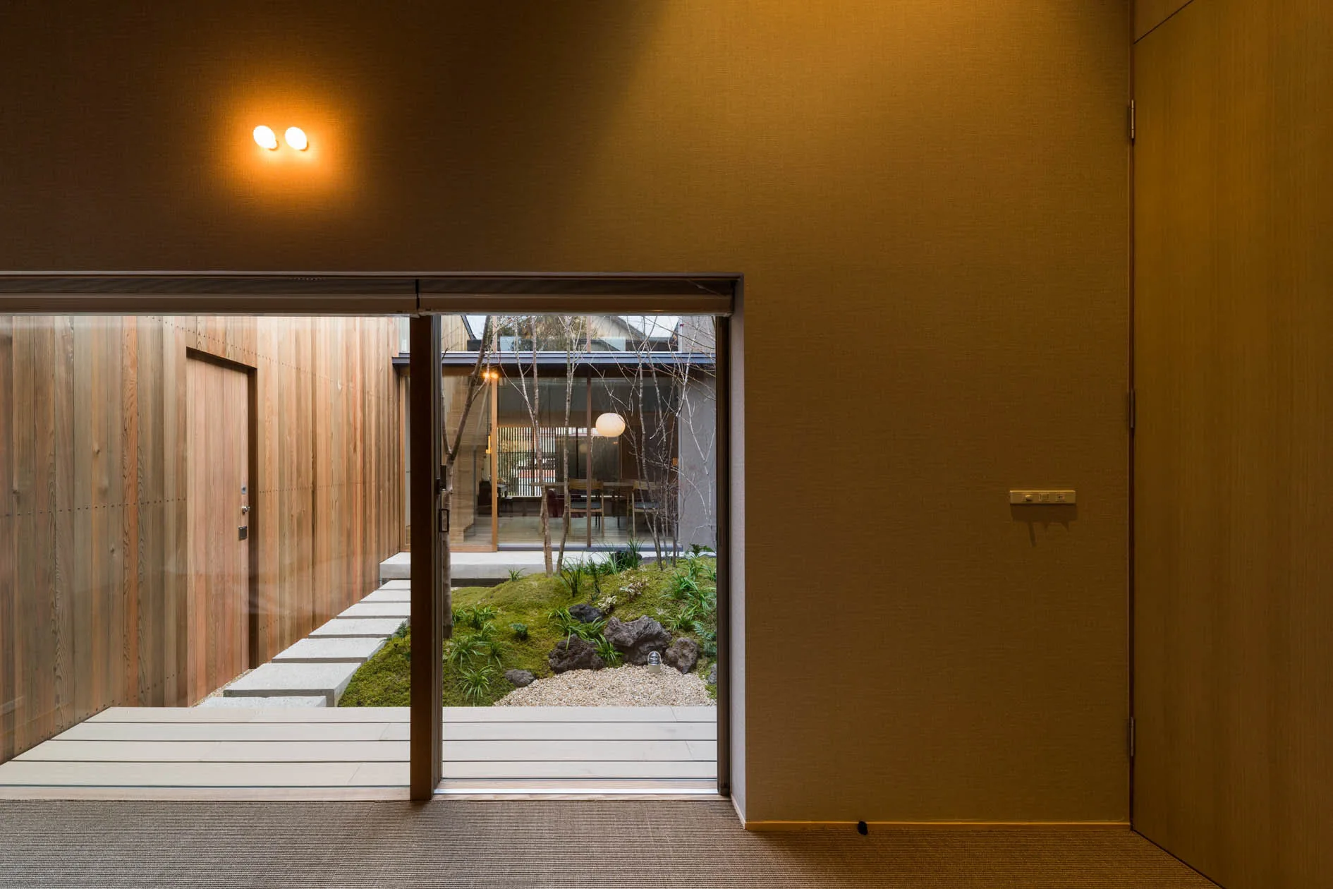 A minimalist room features a large sliding glass door revealing a private courtyard garden with a mossy hill, small trees, and stepping stones.