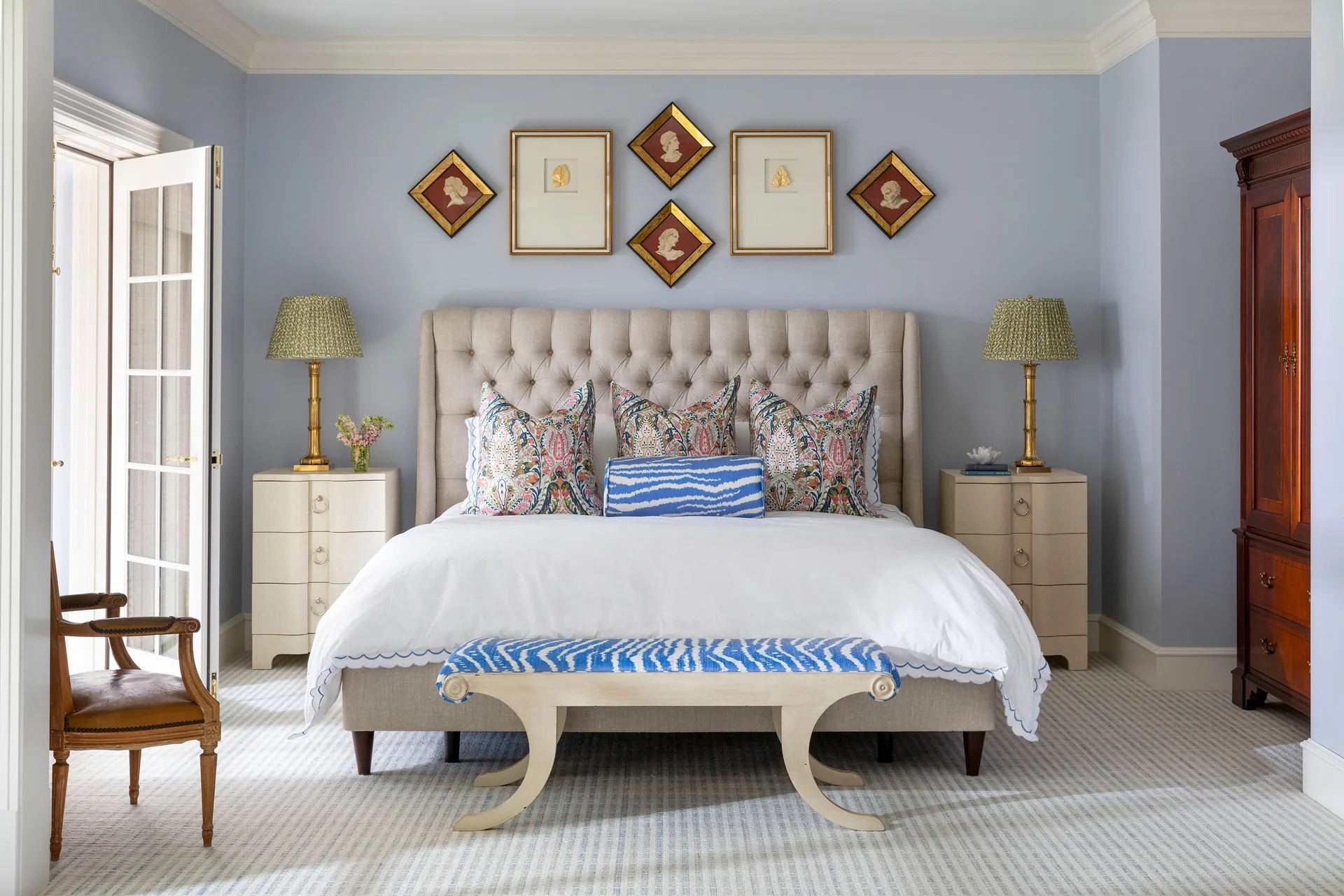 A symmetrical bedroom features a beige tufted headboard, a blue patterned bench, brass lamps, and framed art pieces on a light blue wall.