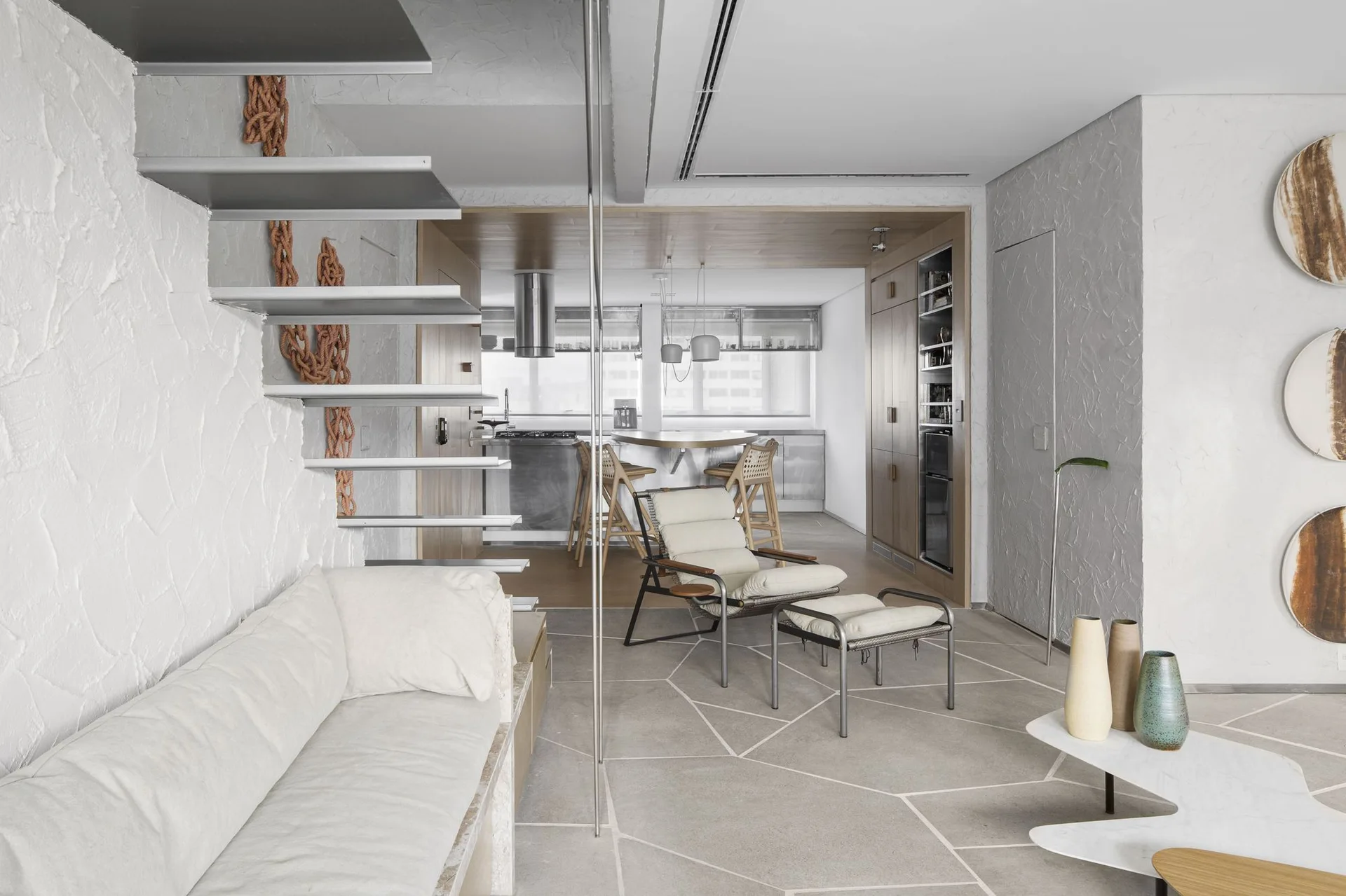 A minimalist living room features a floating metal staircase against a textured white wall, a lounge chair with ottoman, and geometric gray stone tile floors.
