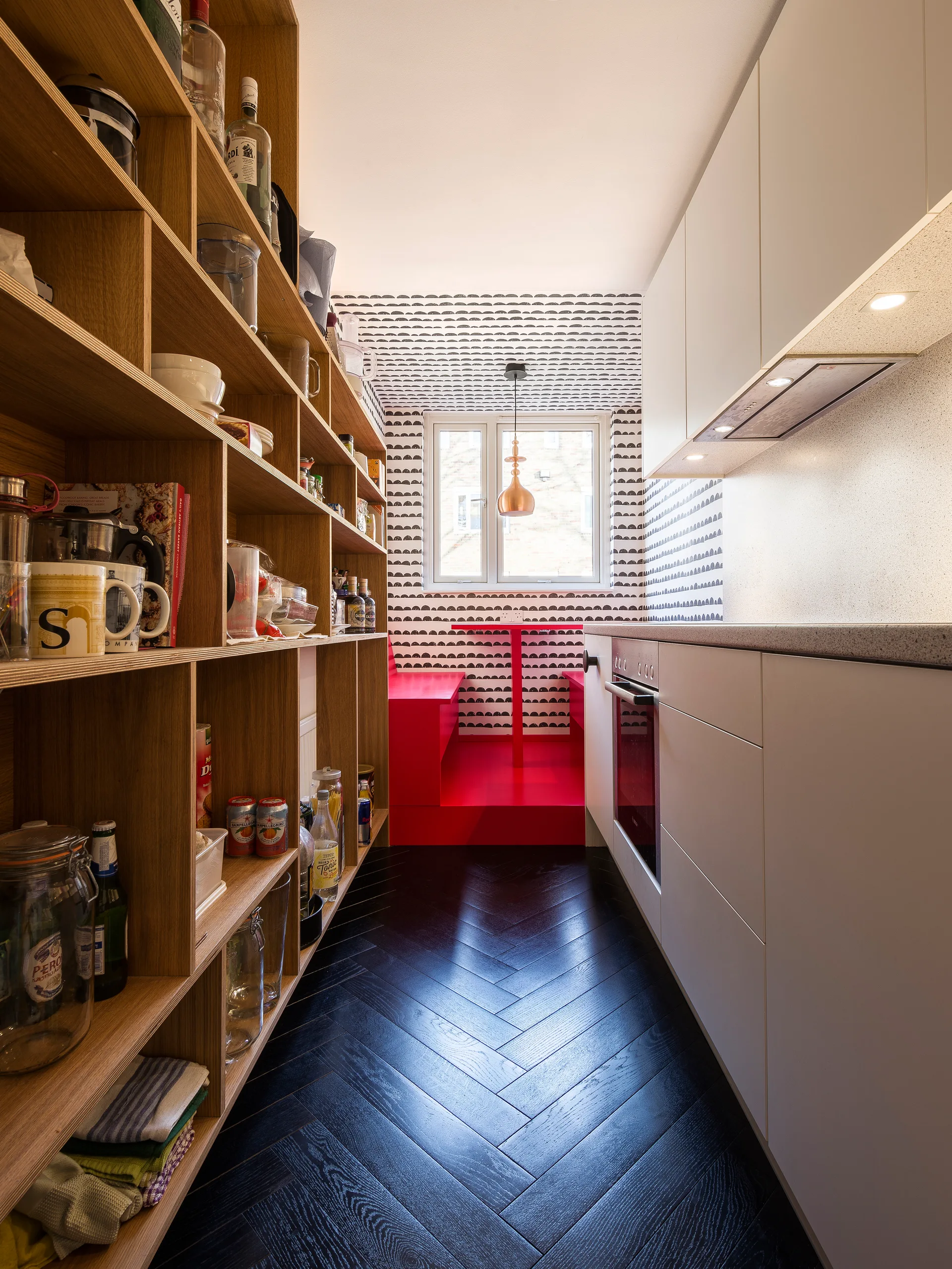 A galley kitchen featuring a full-height wooden shelving unit, white cabinets, and a bright red dining nook at the far end.