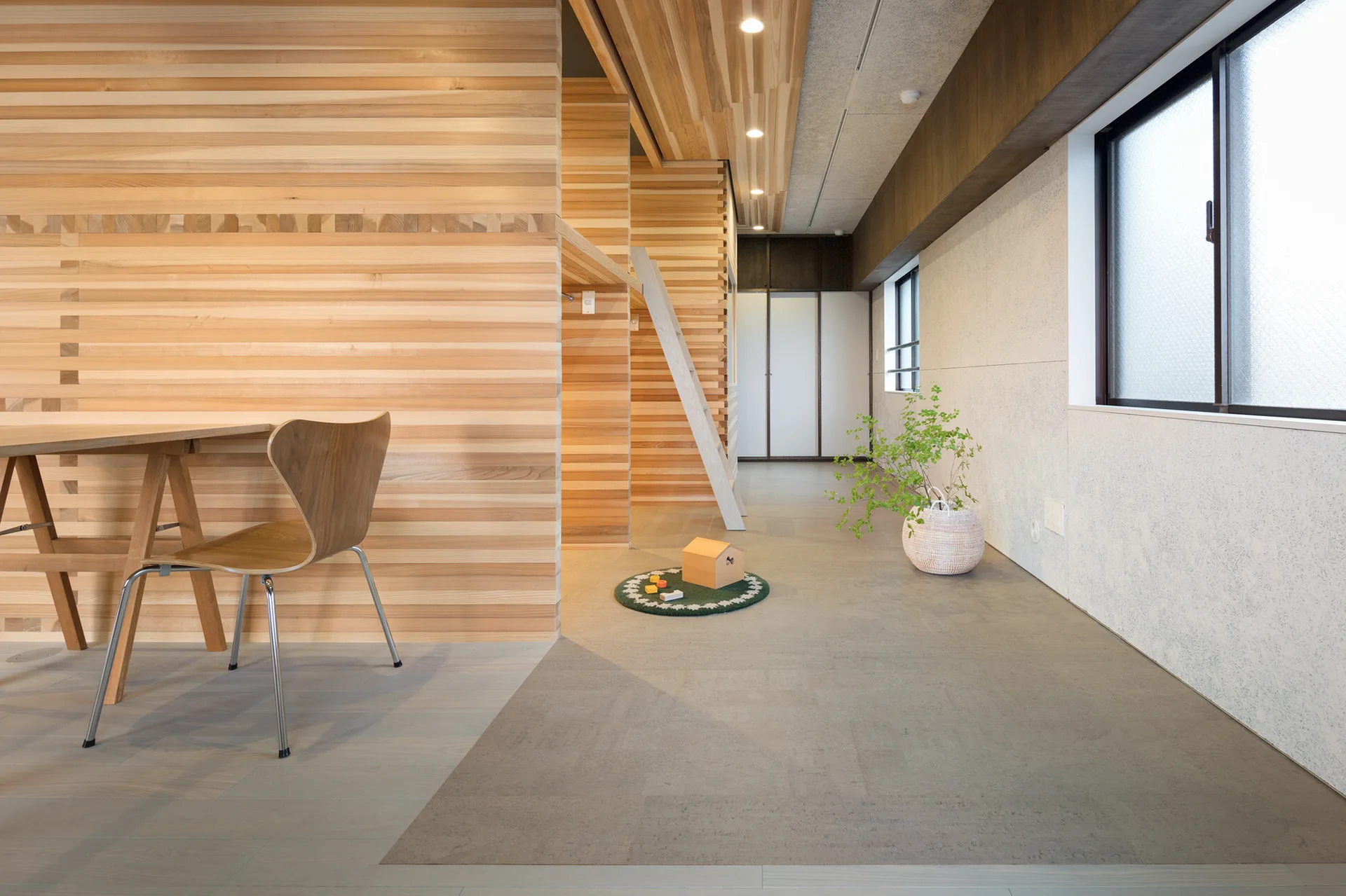 A minimalist living space features light wood horizontal slat paneling on the walls, a wooden desk with a plywood chair, and a grey floor.