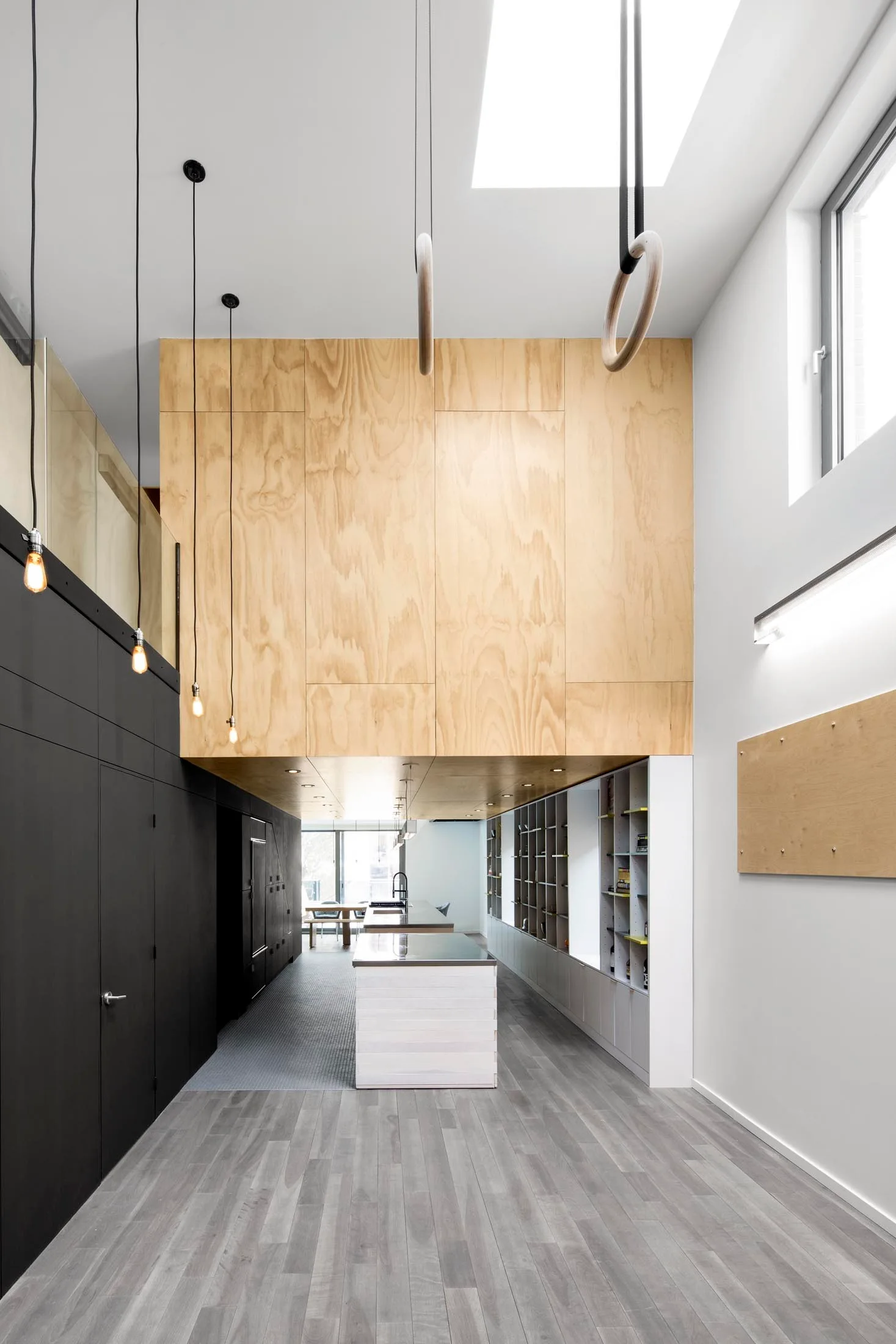 A high-ceiling kitchen with a plywood-clad mezzanine, black cabinetry, a white central island, and wooden rings hanging from a skylight.