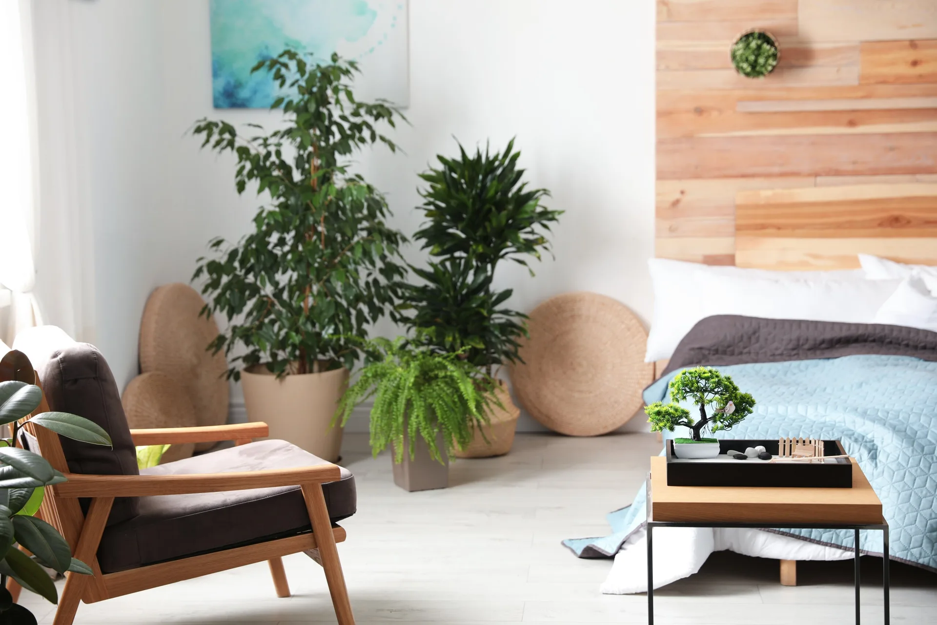 A bright bedroom features a bed with a blue quilt and a wooden headboard, surrounded by potted houseplants, a wooden armchair, and a side table.