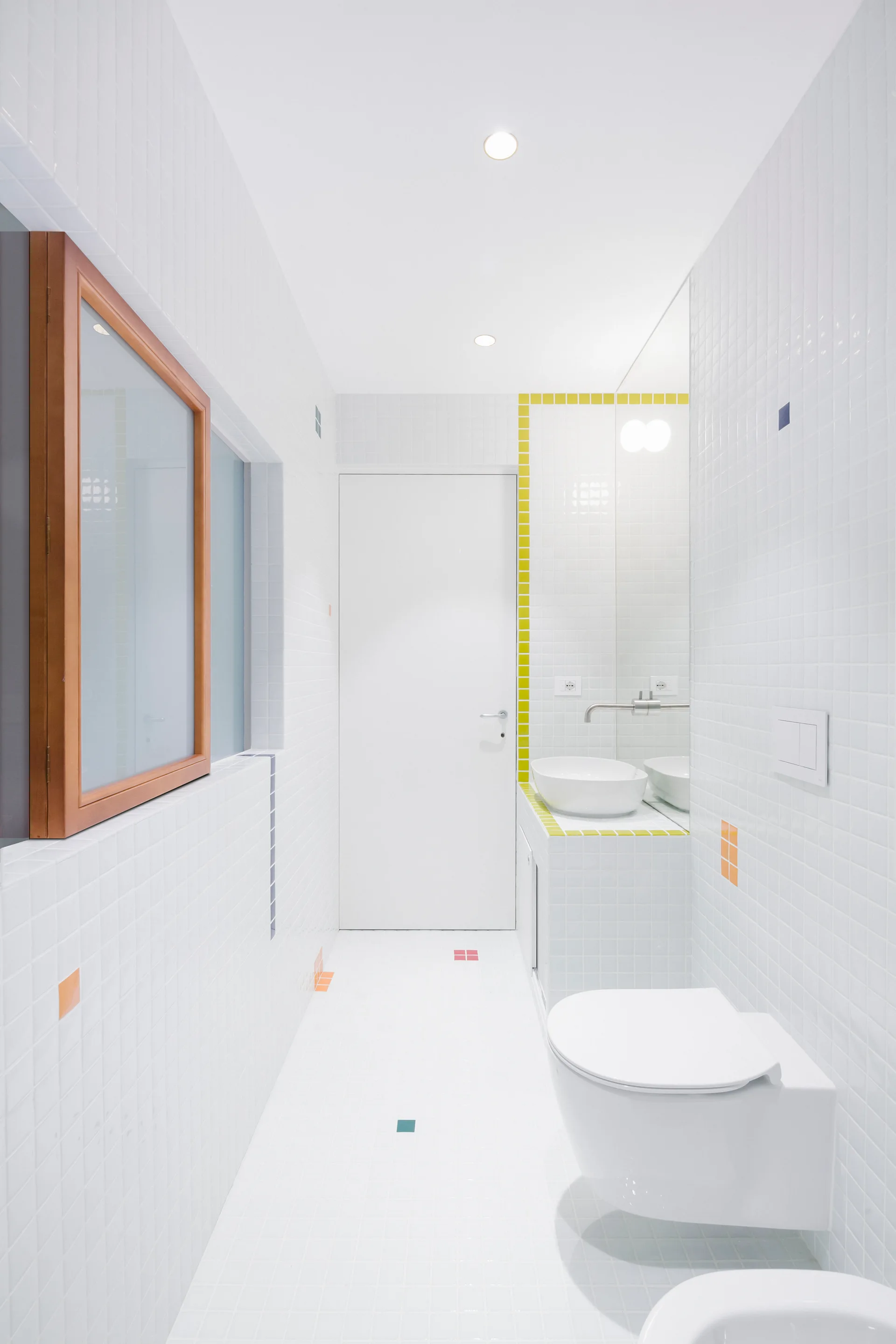 A narrow modern bathroom with white square tiled walls and floor, featuring a wall-hung toilet, a white vessel sink, and a natural wood-framed window.