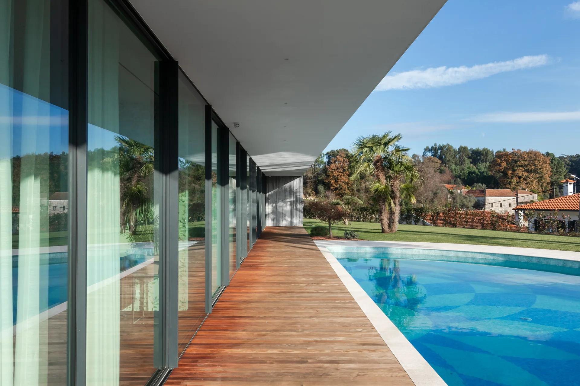 A modern home features a long wood plank deck beneath a flat roof overhang, situated next to a curved blue swimming pool.