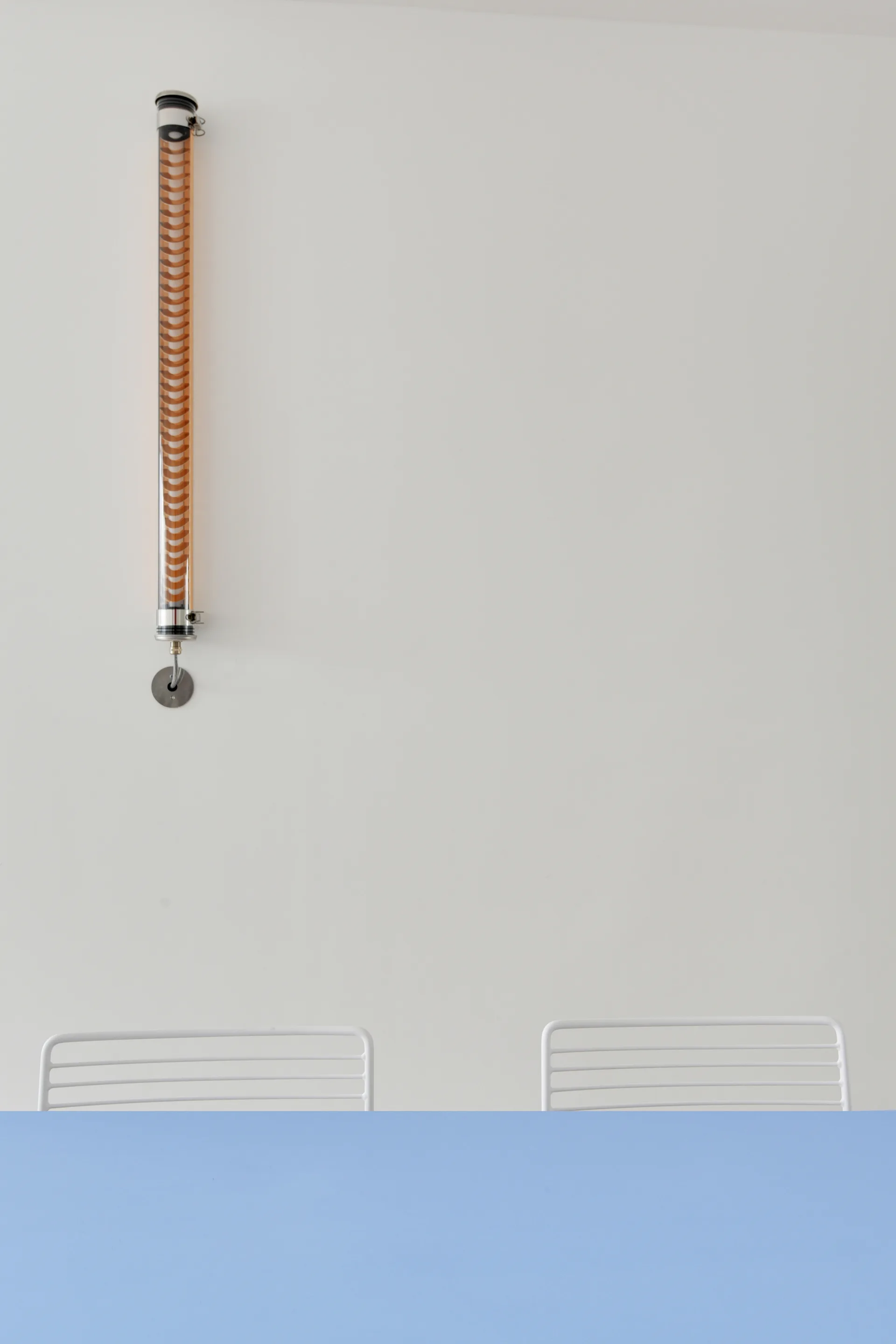An amber cylindrical glass sconce mounted on a plain white wall above white slatted chairs and a light blue tabletop.