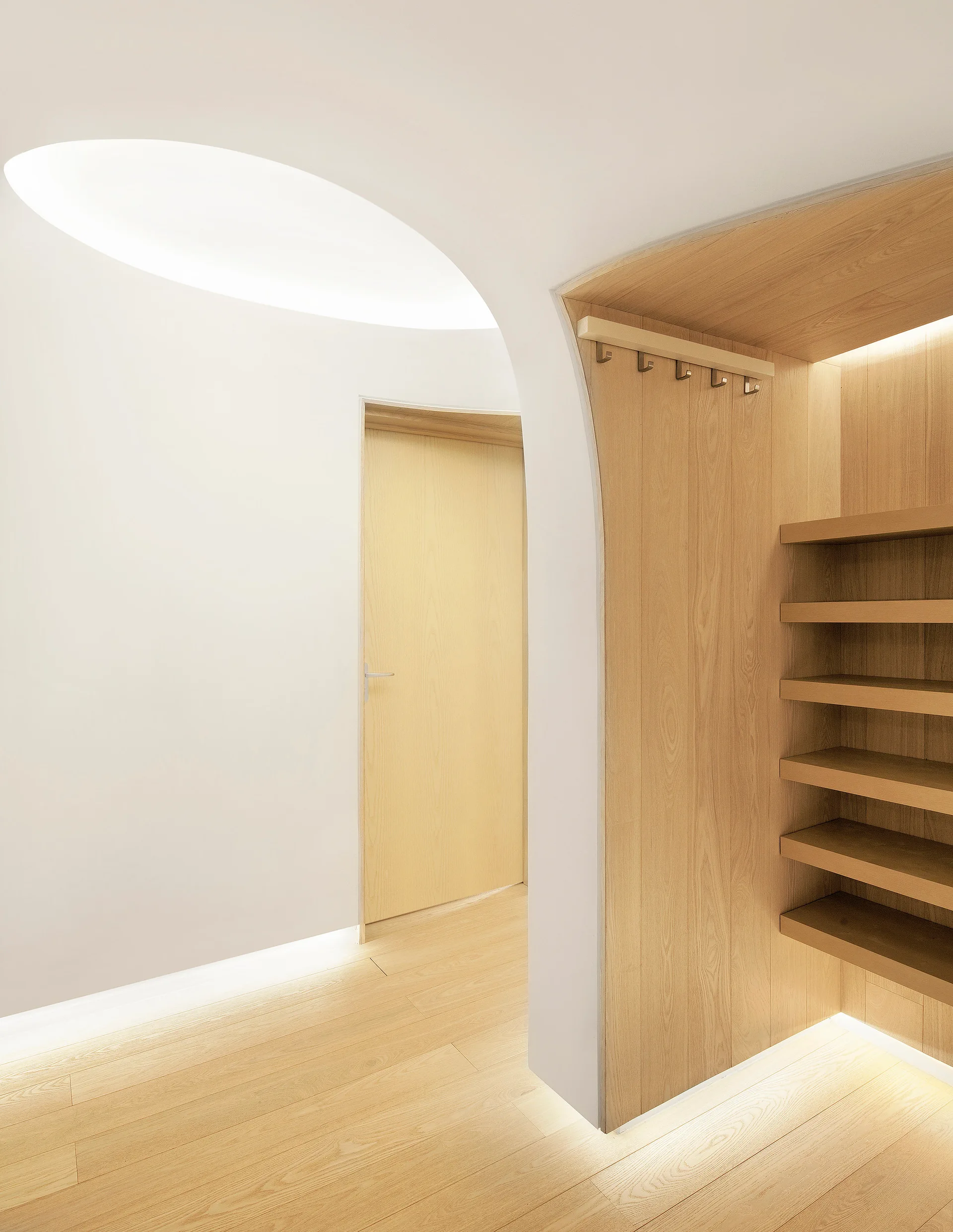 A minimalist white hallway featuring a curved archway, built-in light wood shelving, a matching door, and integrated low-level lighting.