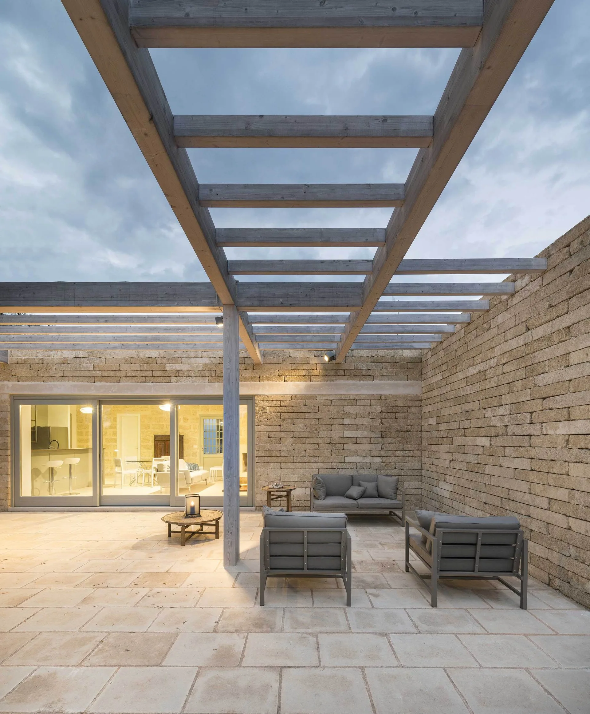 A modern outdoor patio features grey upholstered lounge furniture arranged under a wooden pergola next to a textured stone wall.