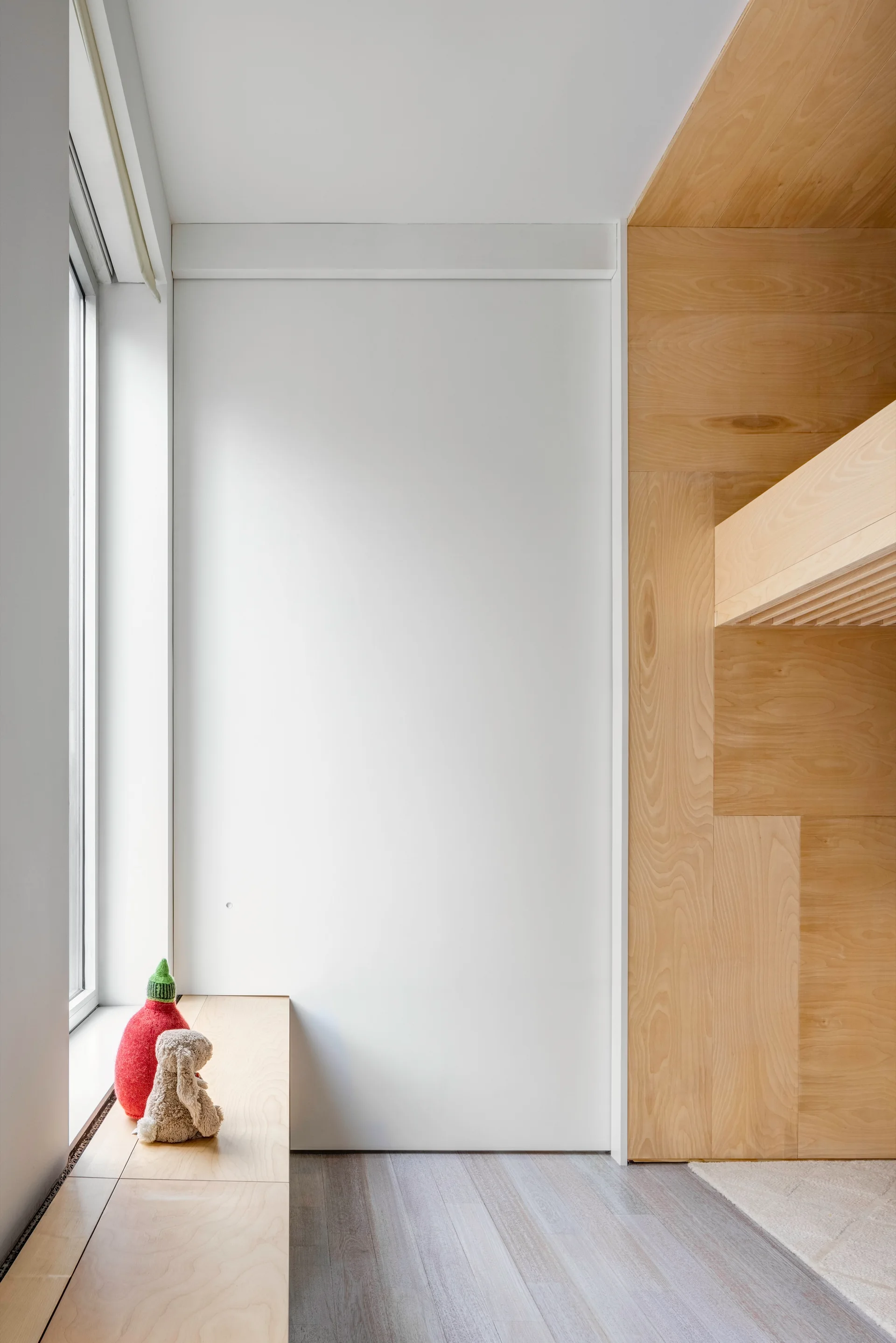 A minimalist room corner features a light plywood built-in bunk bed structure and a low window seat next to a plain white wall.