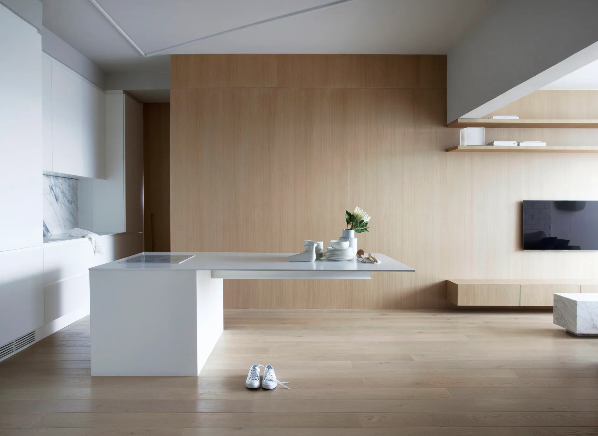 A minimalist kitchen features a white cantilevered island, light wood wall paneling, matching wood floors, and handleless white cabinets.