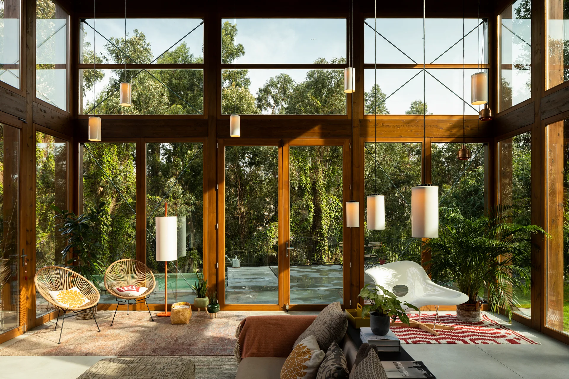 A sunlit living room features floor-to-ceiling wood framed windows overlooking trees, styled with mid-century modern lounge chairs and hanging pendant lights.
