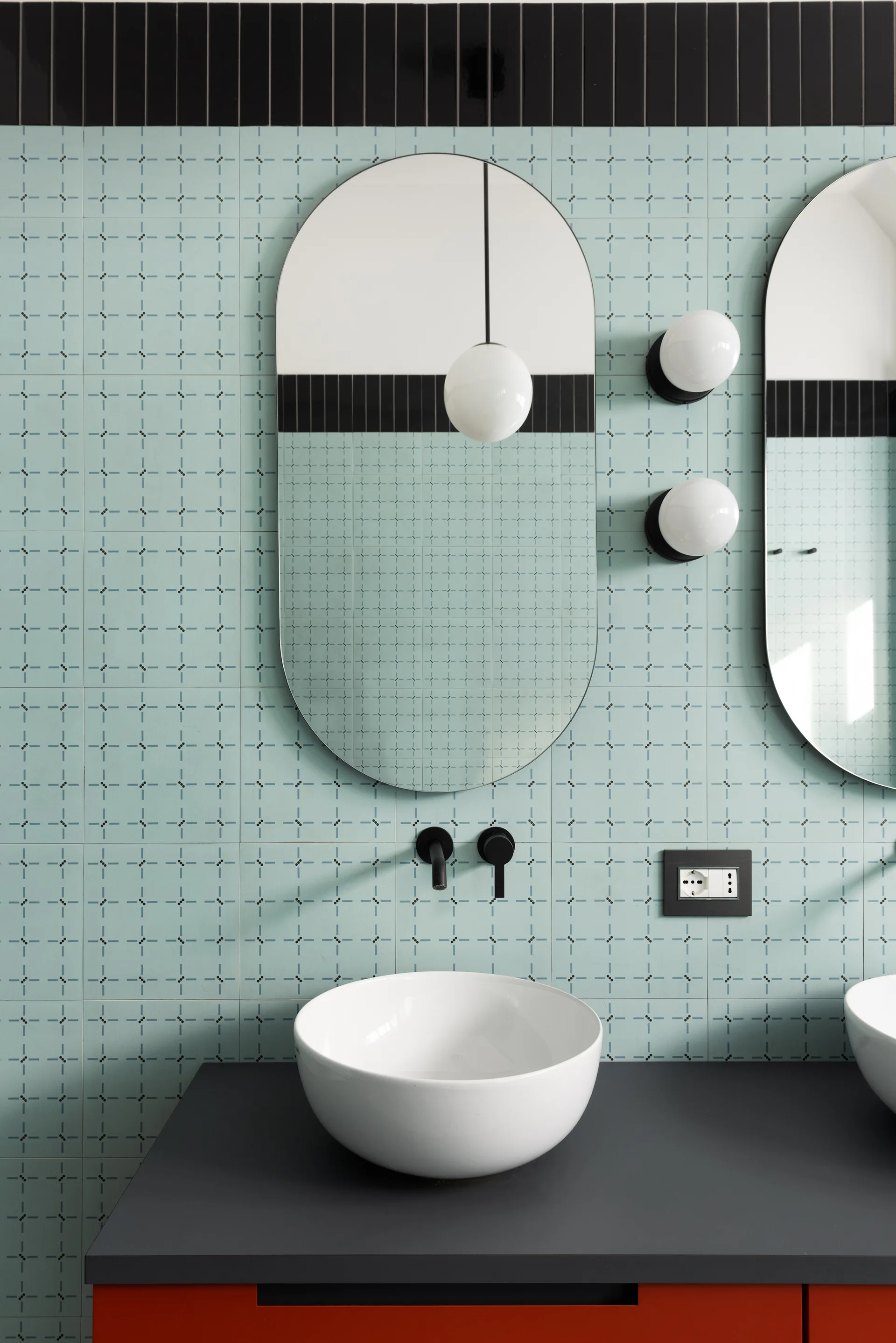 A modern bathroom vanity features a white ceramic vessel sink, a matte black countertop, oval mirrors, and light blue patterned wall tile.