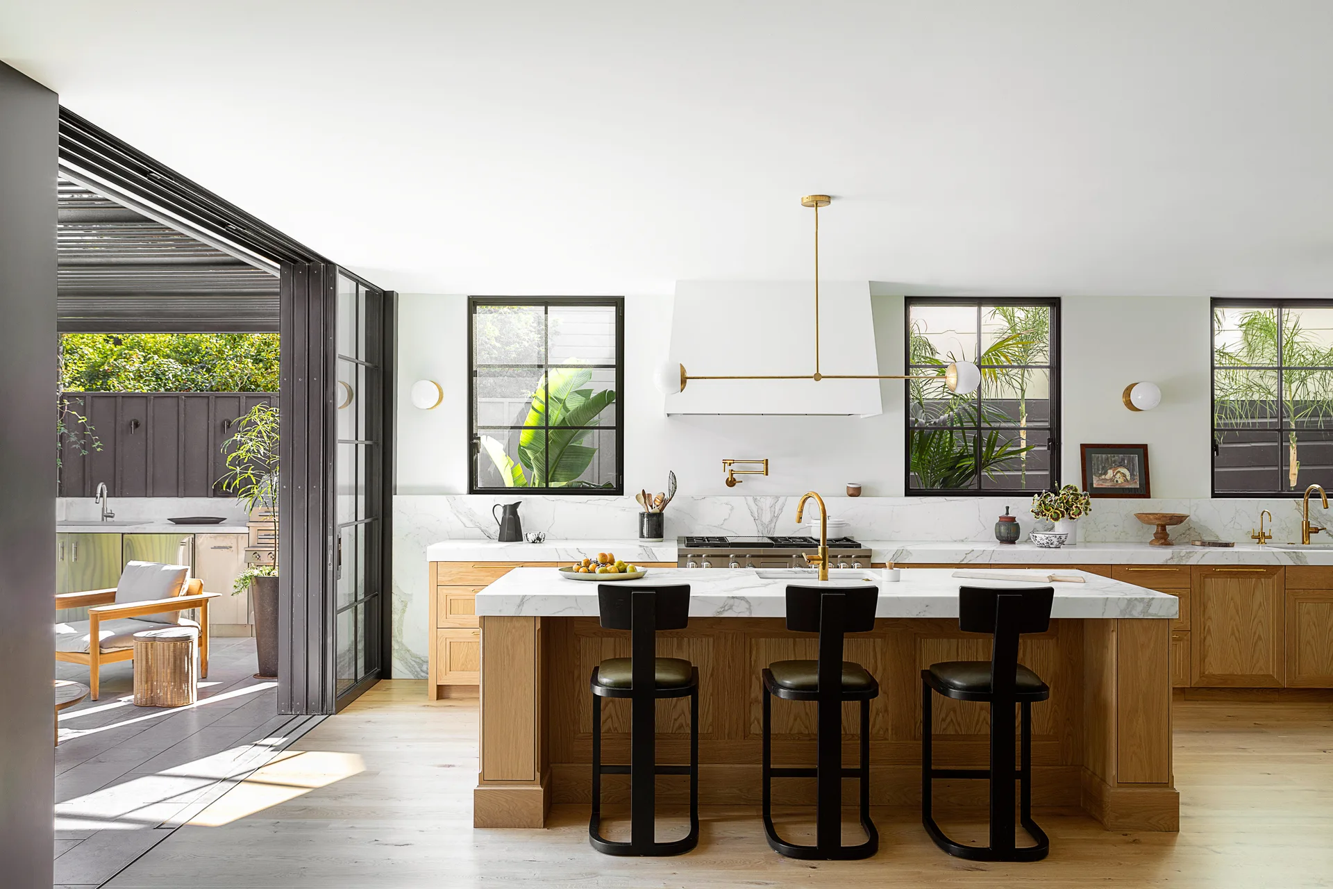 A bright modern kitchen features a wood-toned island with marble countertops, black bar stools, and large sliding glass doors leading to a patio.