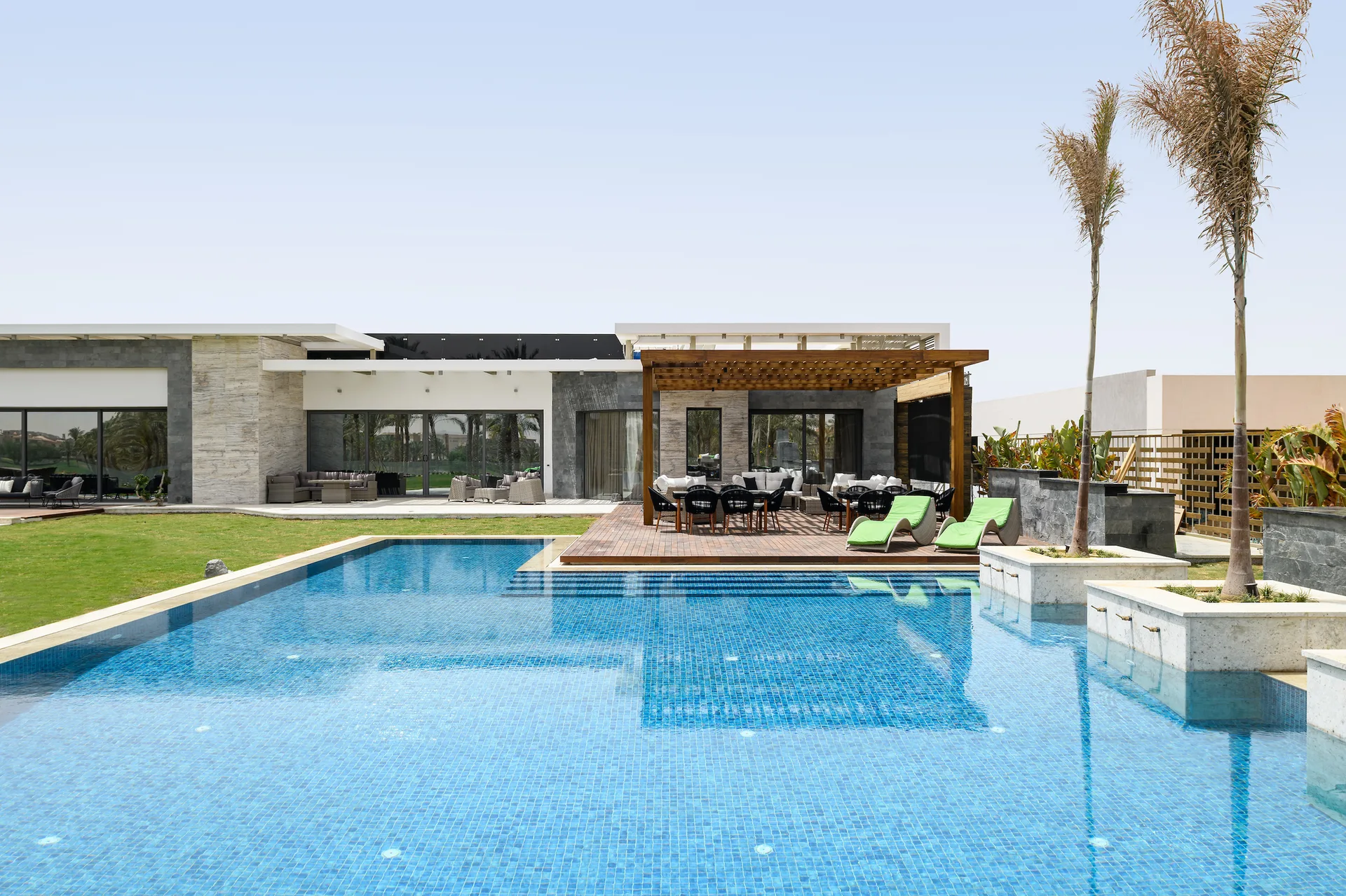 A large blue tiled swimming pool sits next to a wooden patio deck with a dining table under a pergola and two green lounge chairs.