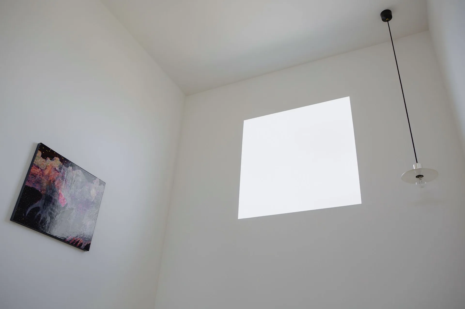 A minimalist white wall with a square window, a thin black pendant light, and a dark framed painting.