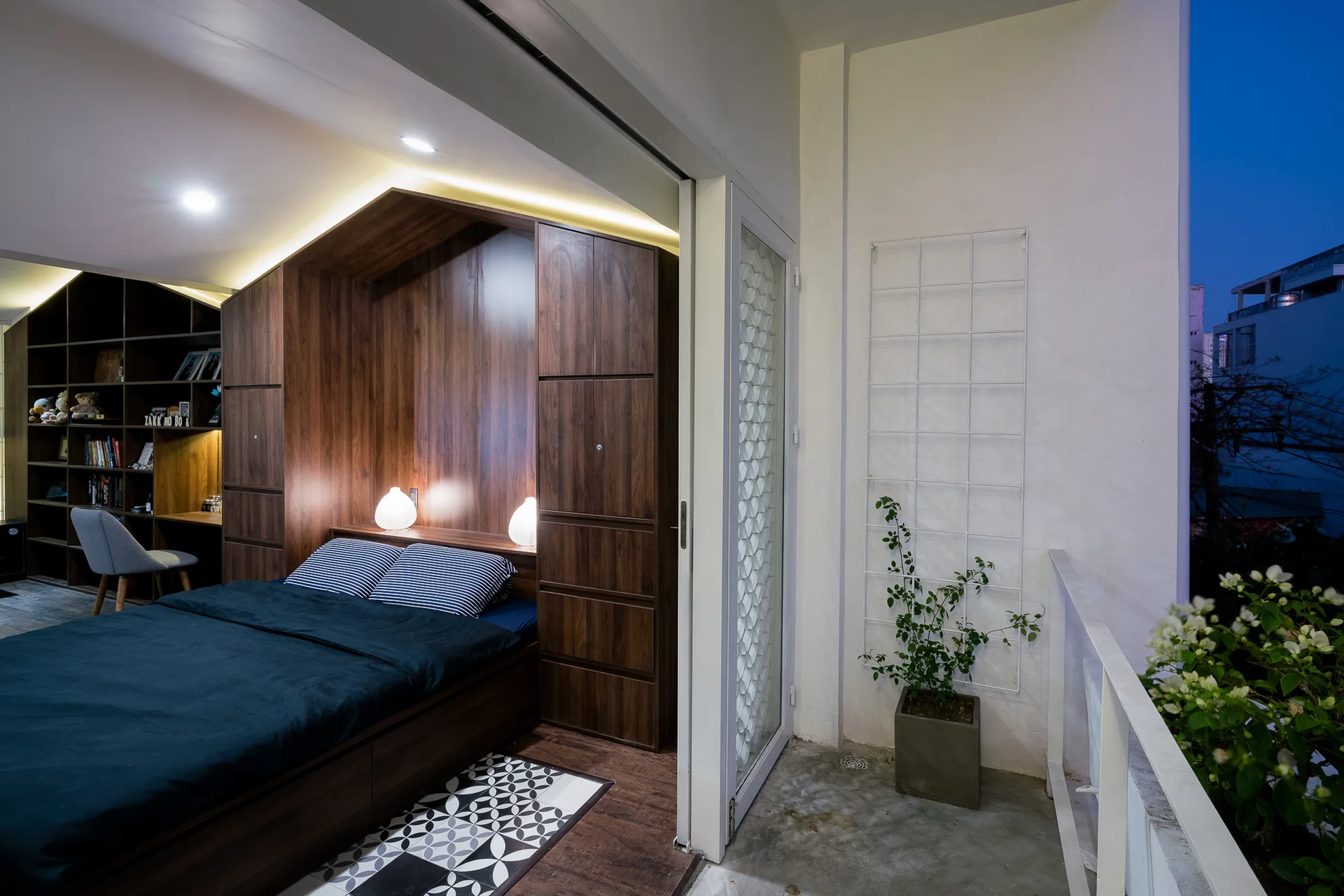 A modern bedroom features a built-in dark wood wardrobe framing a bed, next to a sliding glass door that opens to a balcony.