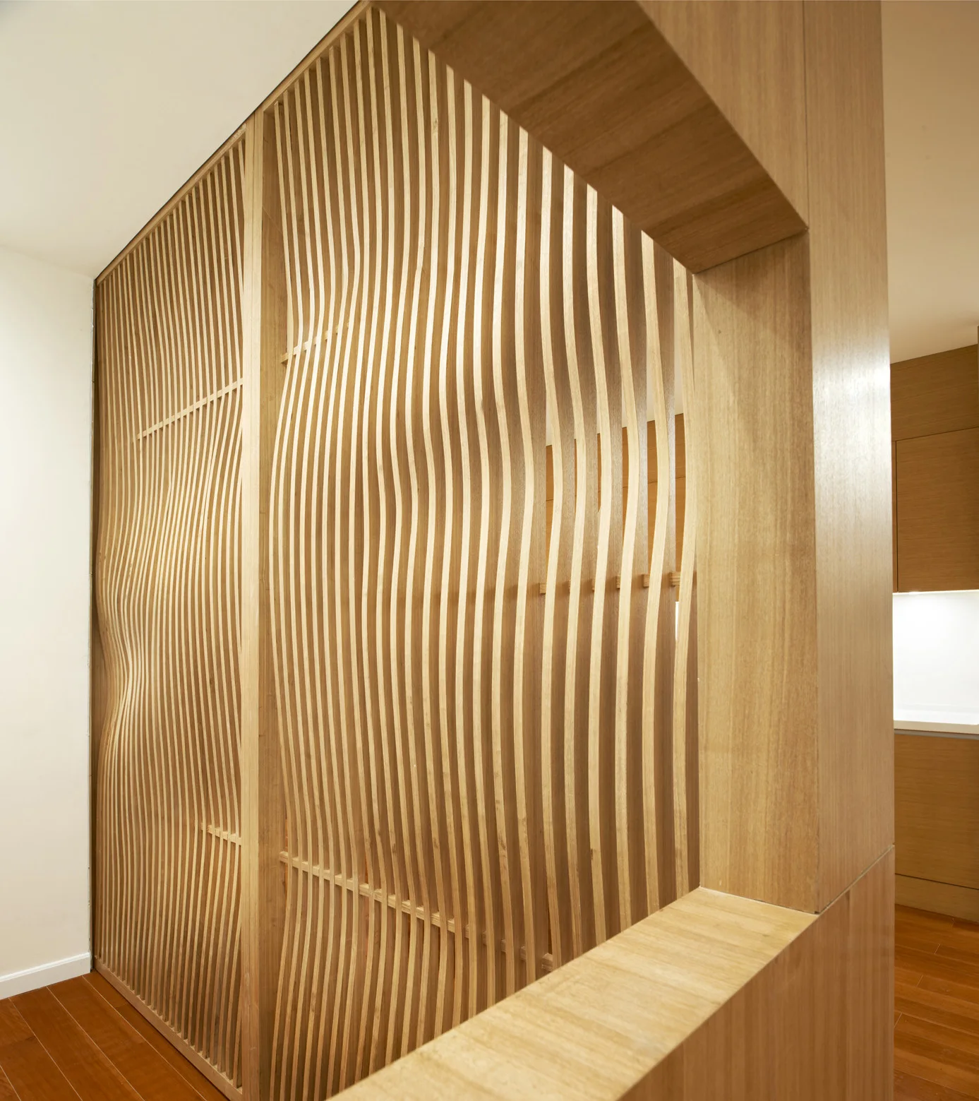 A custom room divider constructed from vertical wavy wooden slats stands beside a smooth white wall and warm wood flooring.