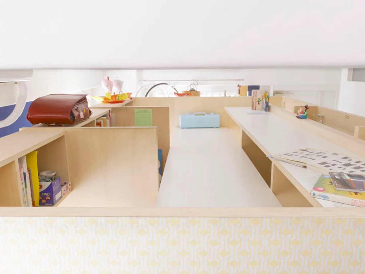 A high-angle view of a custom light wood loft study area featuring integrated desks, storage compartments, books, and a yellow patterned front panel.