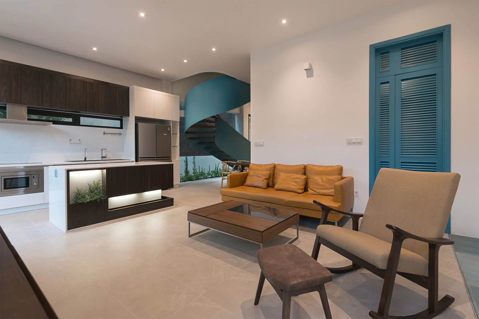A modern open-concept living room and kitchen featuring a tan leather sofa, a wooden rocking chair, and a teal spiral staircase in the background.