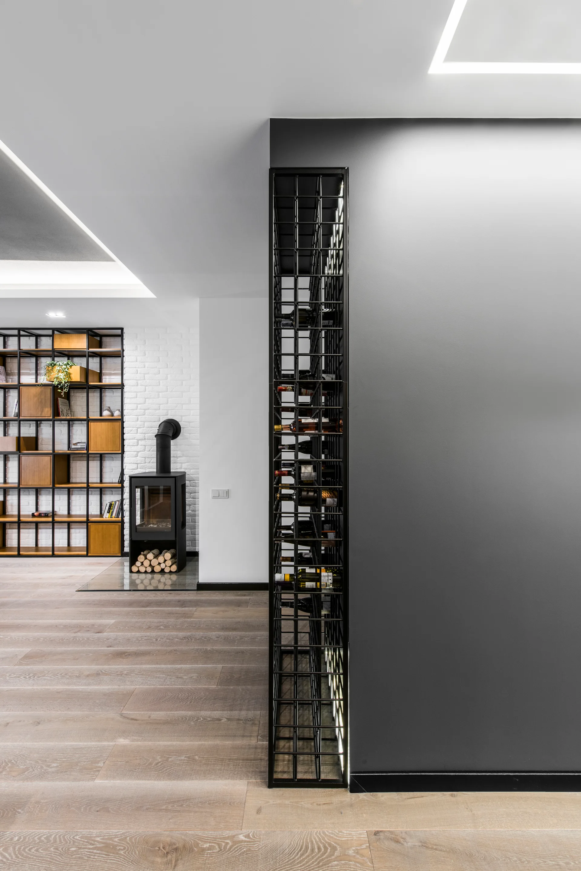 A modern interior space featuring a tall vertical black metal wine rack built into a grey partition wall next to a wood-burning fireplace.