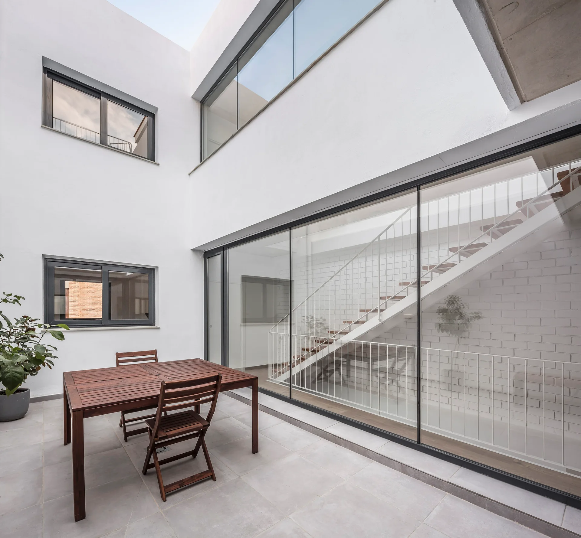 A minimalist outdoor courtyard features a wooden dining set on light grey tiles next to a large glass wall showing an interior staircase.