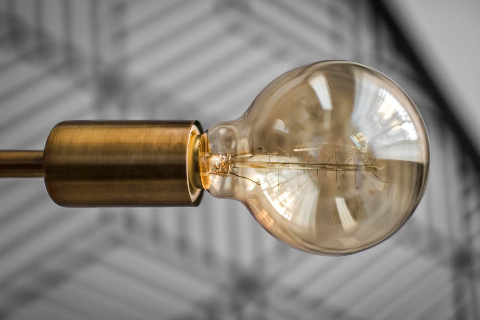 A close-up of a round amber glass filament light bulb secured in a brushed brass metallic socket against a blurred patterned background.
