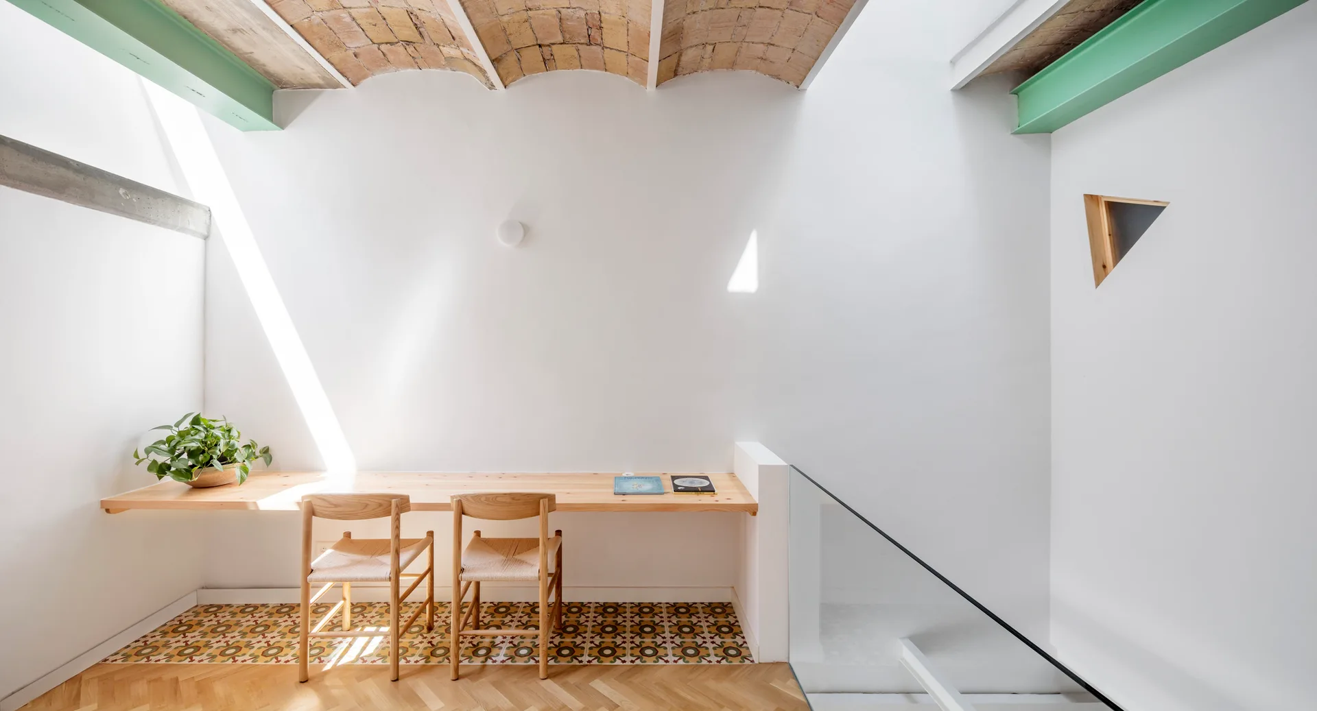A minimalist workspace features a floating wood desk with two chairs beneath an exposed vaulted brick ceiling and a bright skylight.