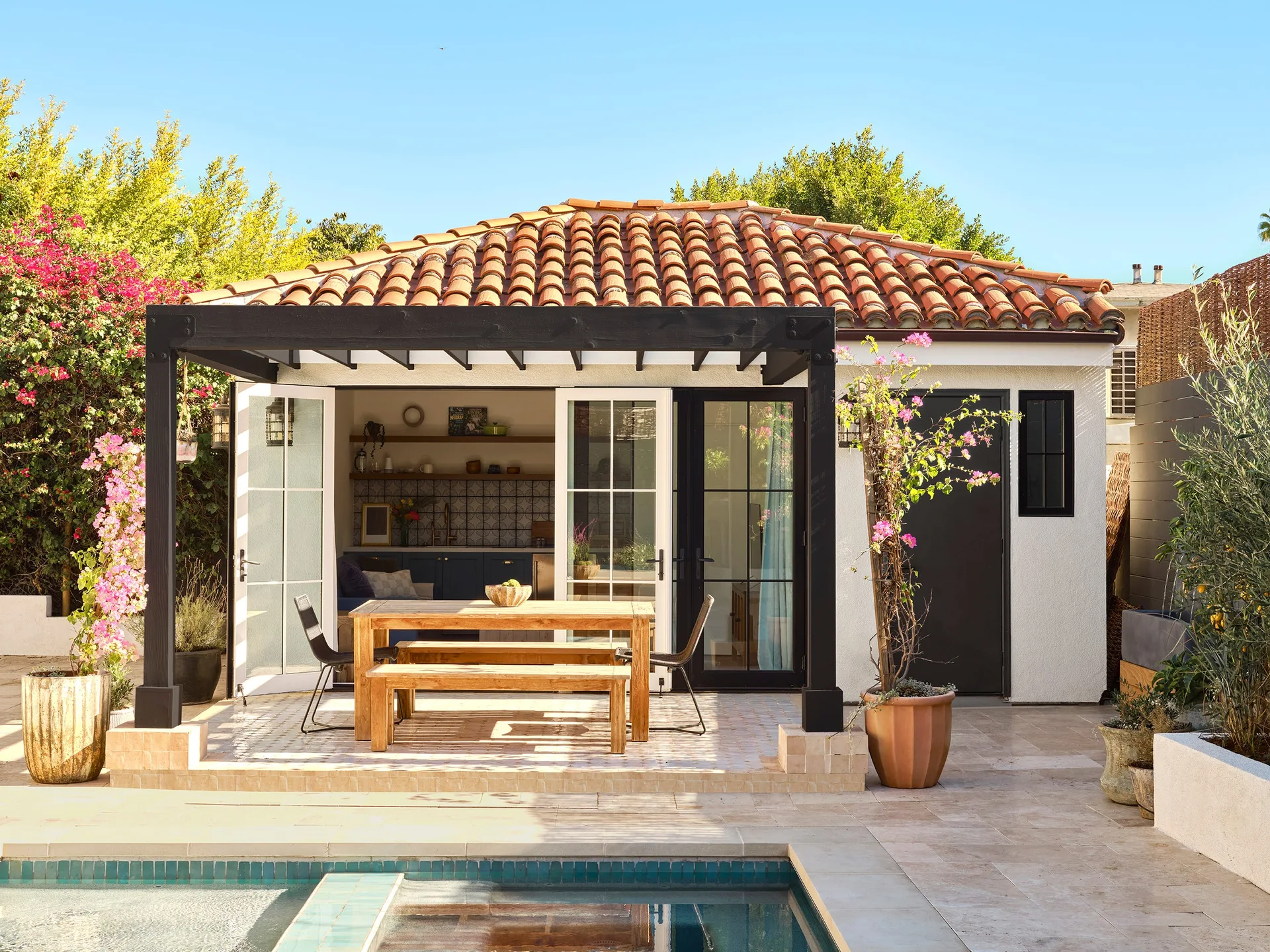 A sunny poolside patio features a wooden dining set under a black pergola next to a white stucco casita with a terracotta tile roof.