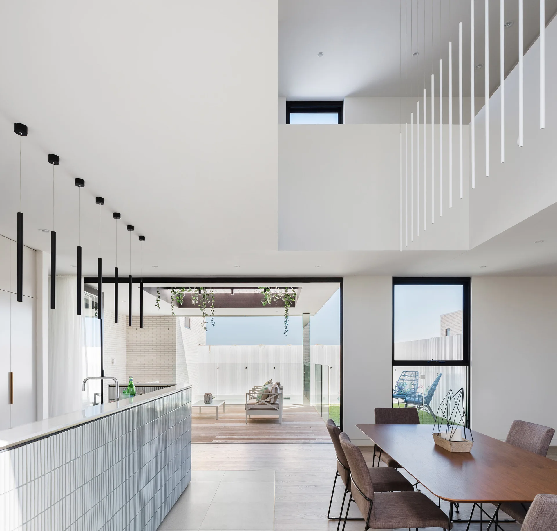 An open-concept modern kitchen features a grey ribbed tile island, black pendant lights, a wood dining table, and large glass doors opening to a patio.