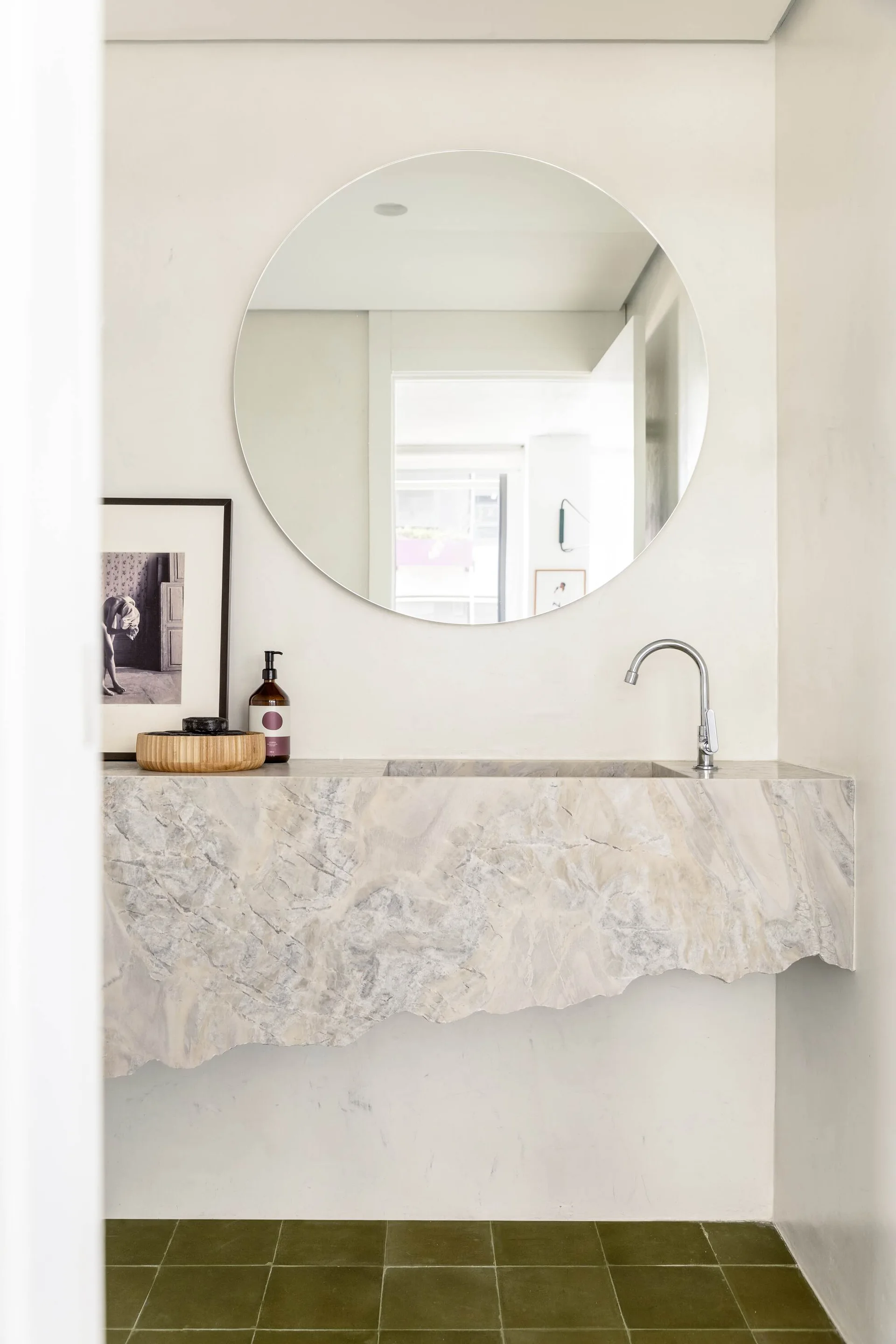 A minimalist bathroom features a floating stone vanity with a raw chiseled edge, a round mirror, and olive green floor tiles.