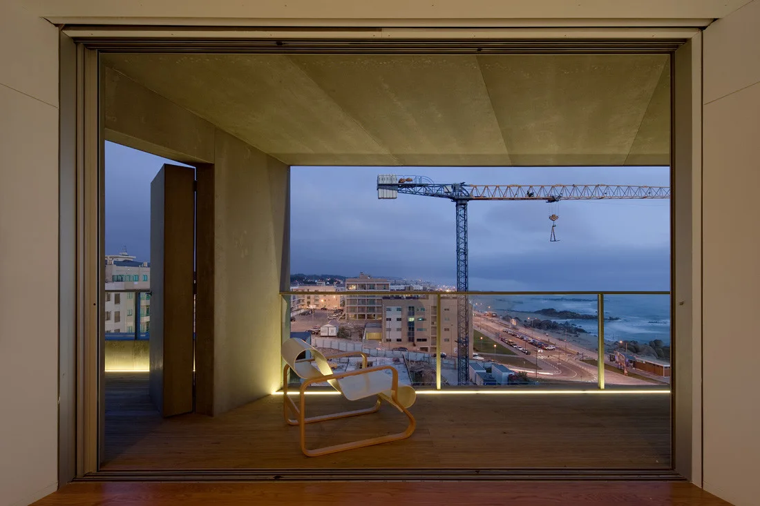 A minimalist concrete balcony at twilight features a curved wood armchair, wood plank decking with linear floor lighting, and a coastal city view.