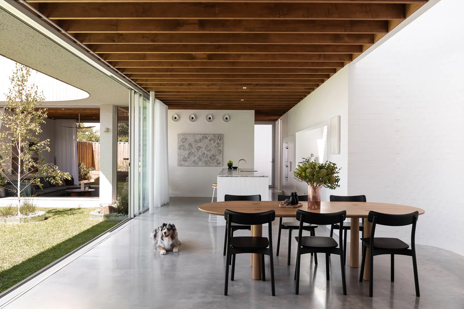 An open dining area features an oval wood table with black chairs on polished concrete floors under an exposed timber beam ceiling.