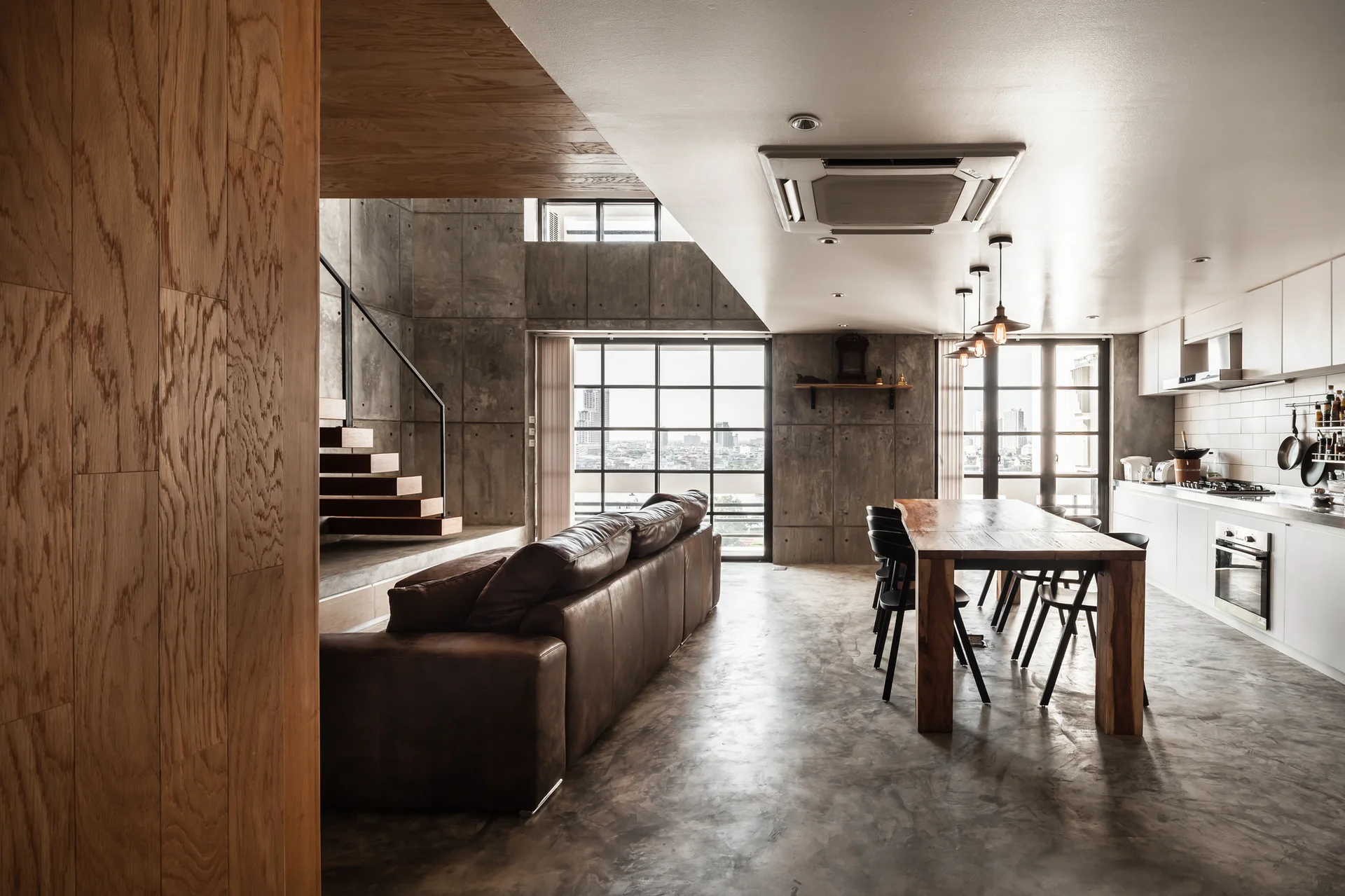 An industrial open-plan living space features a brown leather sofa, a rustic wooden dining table, concrete paneled walls, and metal grid windows.